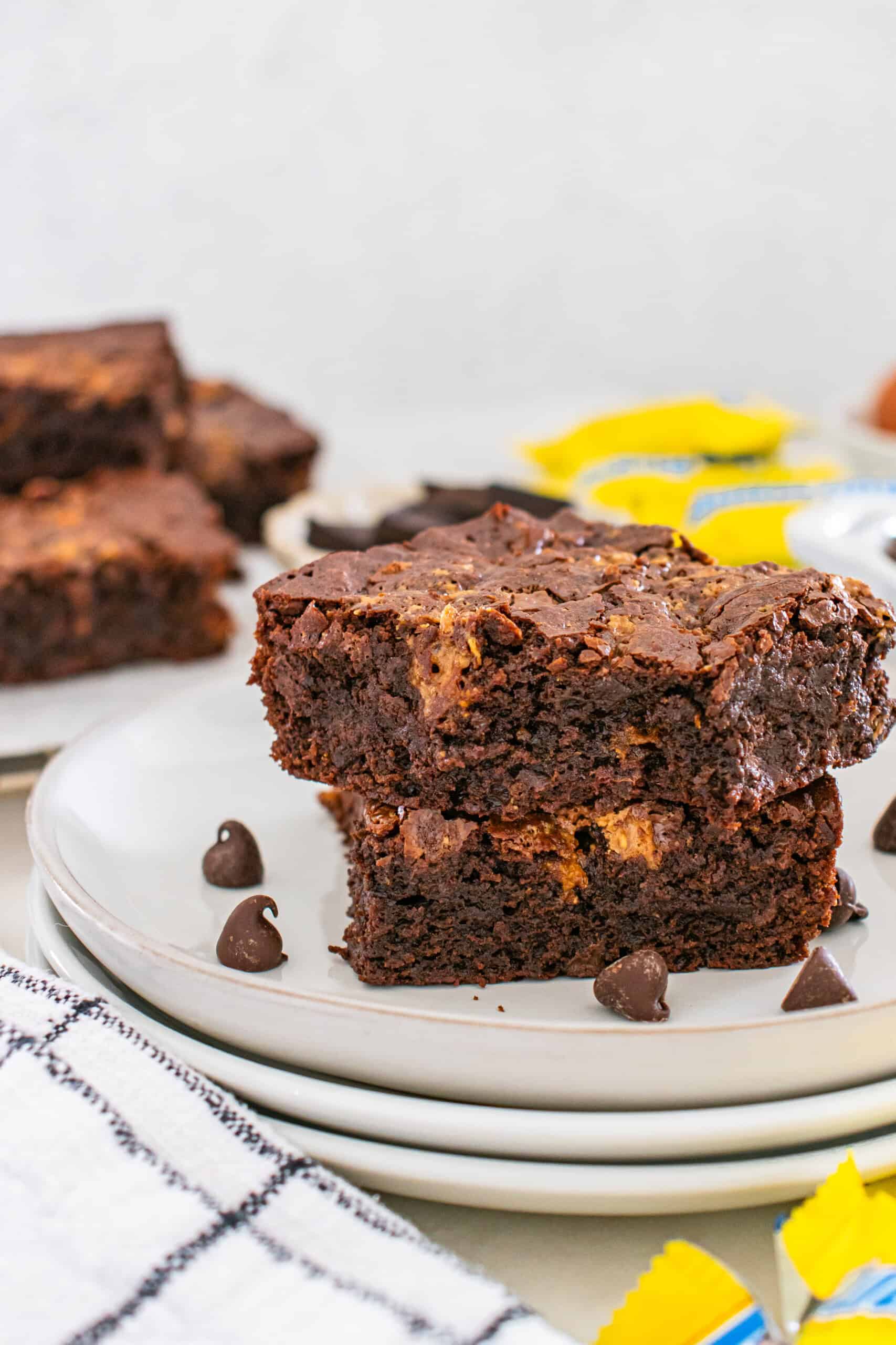 Two chocolate brownies with a marbled texture are stacked on a white plate, with chocolate chips scattered nearby and additional brownies in the background.