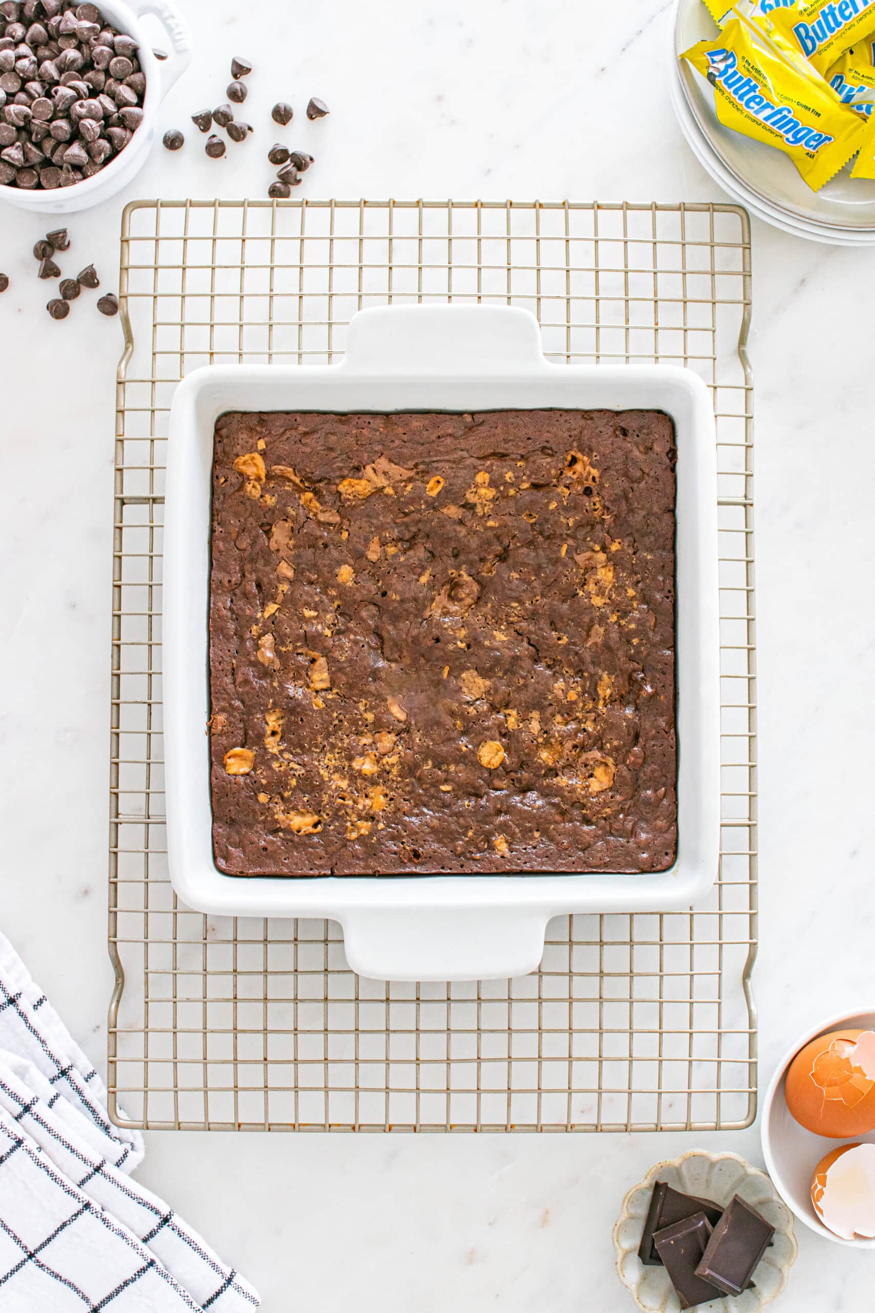 Square chocolate brownie slab in a white baking dish on a cooling rack, surrounded by chocolate chips, butter, eggs, and a kitchen towel on a marble surface.
