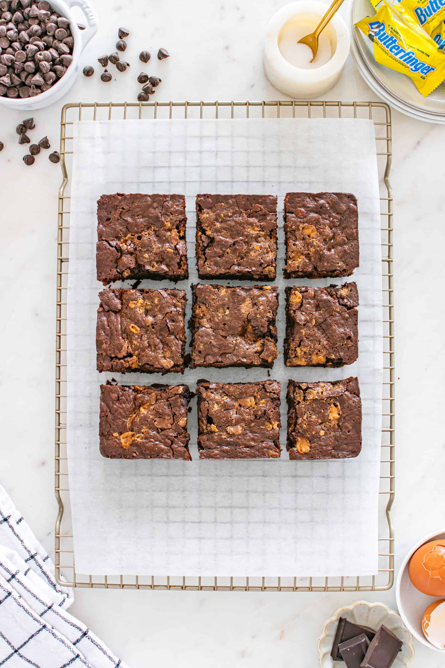 Nine chocolate brownies with visible caramel or candy pieces are arranged in a 3x3 grid on a wire rack lined with parchment paper, surrounded by baking ingredients.
