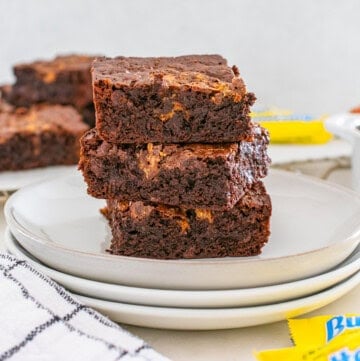 Three chocolate brownies stacked on two white plates, with more brownies, a bowl of chocolate chips, and yellow candy wrappers in the background.