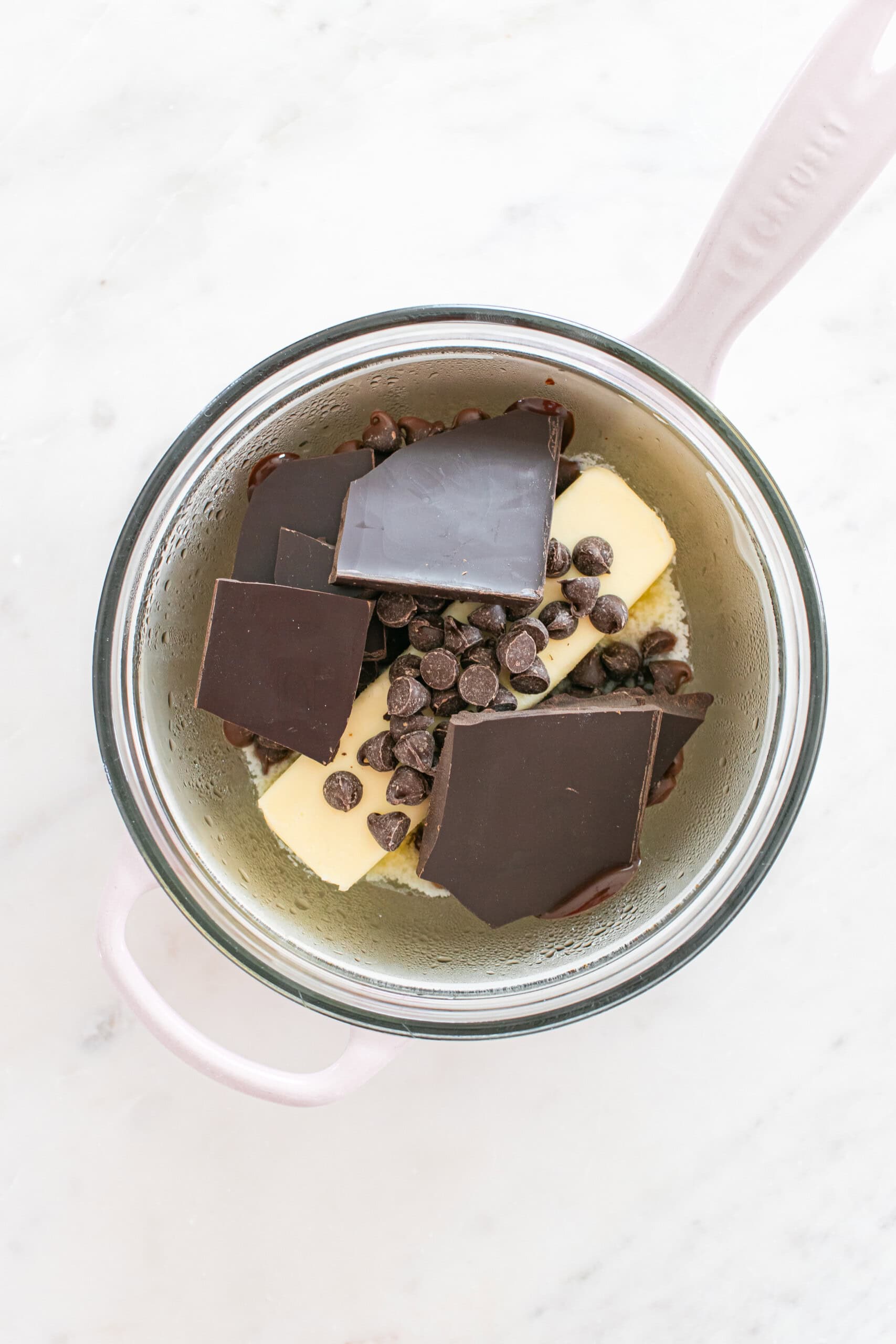 A glass bowl with butter, chocolate chunks, and chocolate chips sits over a pot, preparing ingredients to melt on a white marble surface.