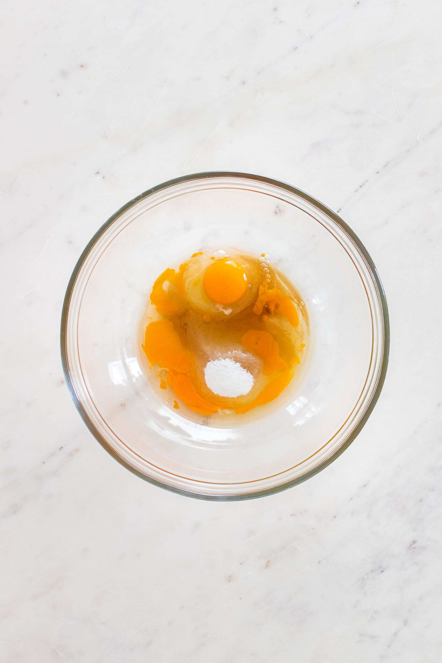 A glass bowl containing cracked raw eggs and a small amount of white granulated sugar on a white marble surface.