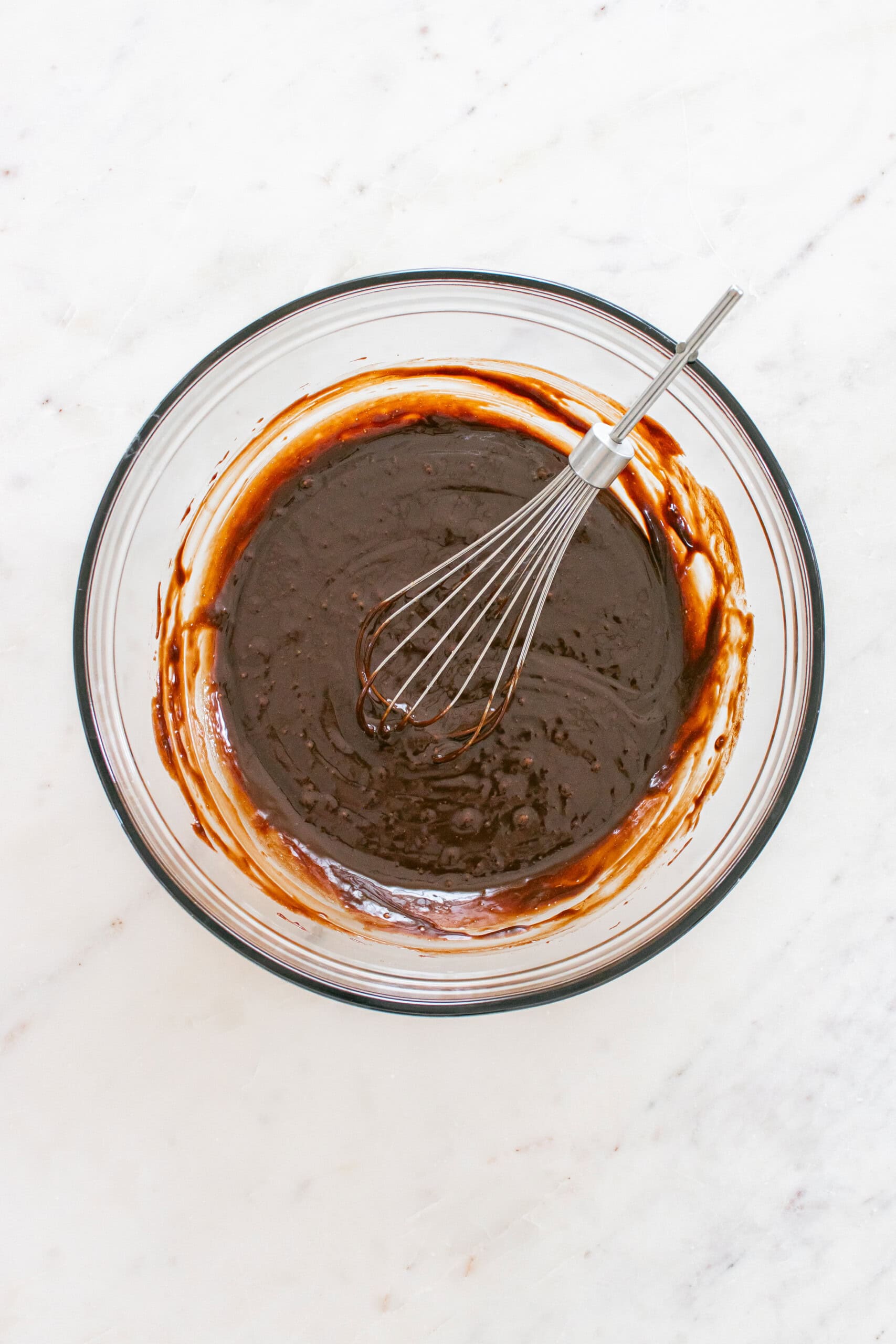 A glass bowl containing chocolate batter with a metal whisk resting inside, placed on a white marble surface.