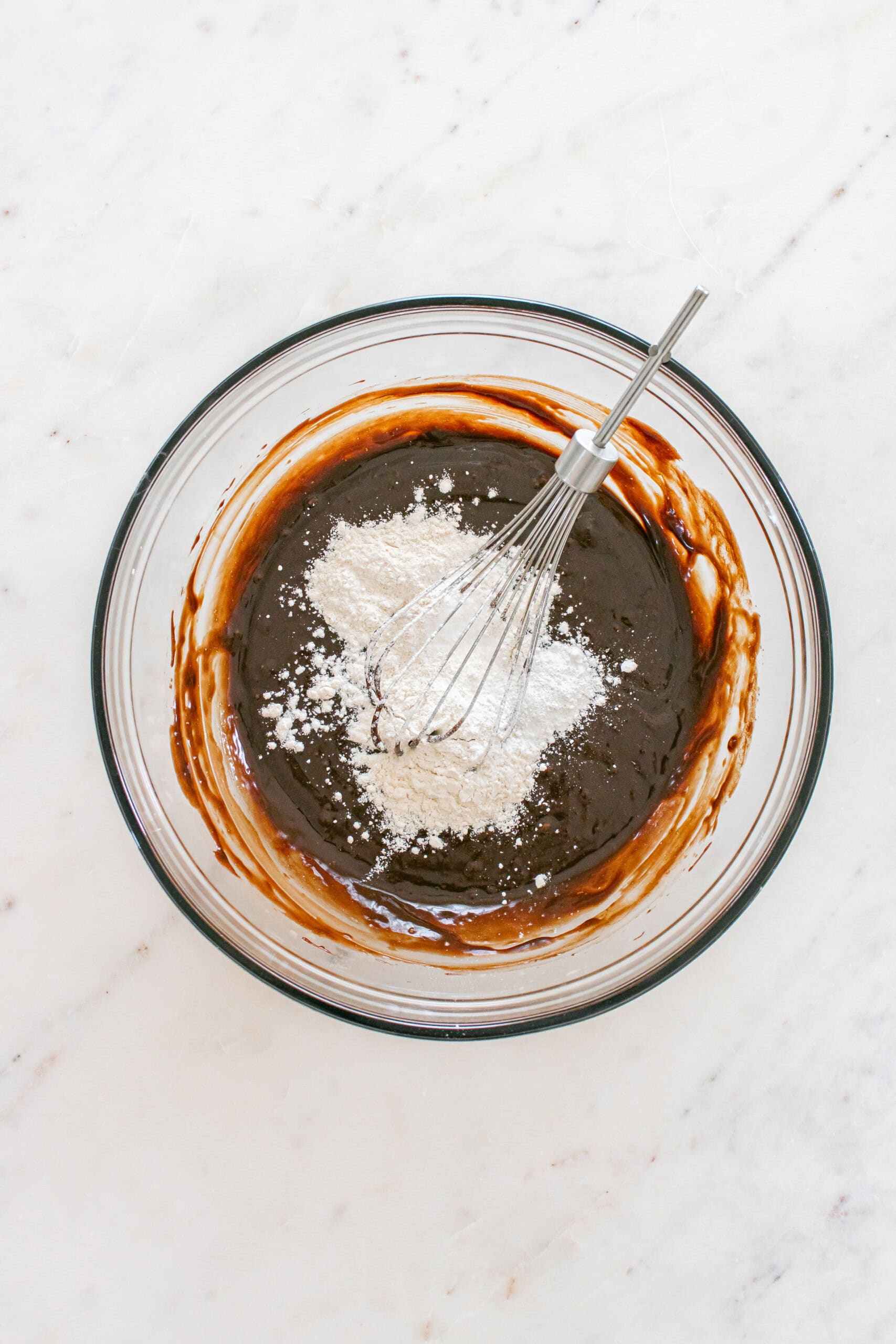A glass bowl with chocolate batter and a whisk, with flour being mixed in, on a white marble surface.