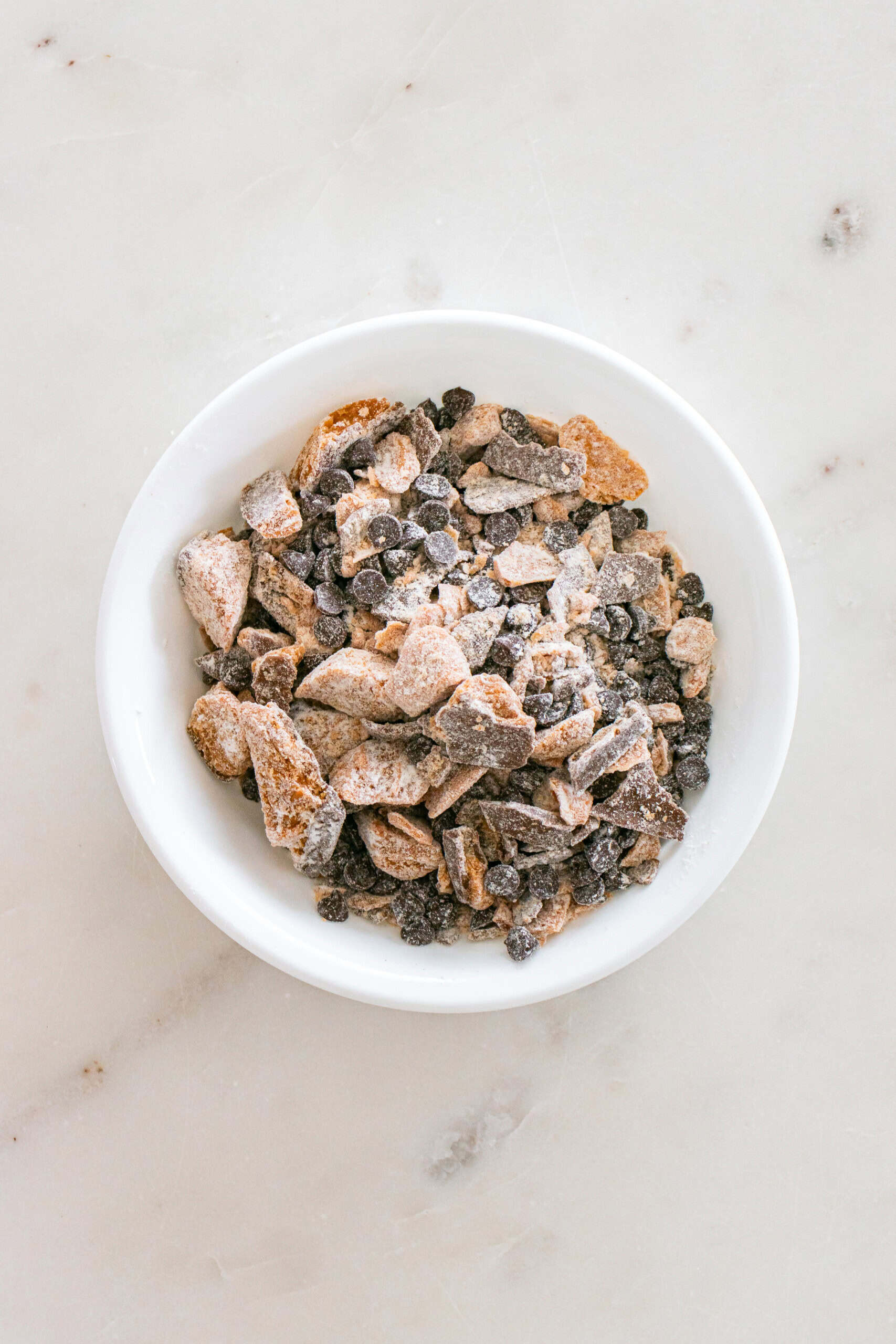 A white bowl filled with assorted chocolate and toffee chunks sits on a light marble surface.
