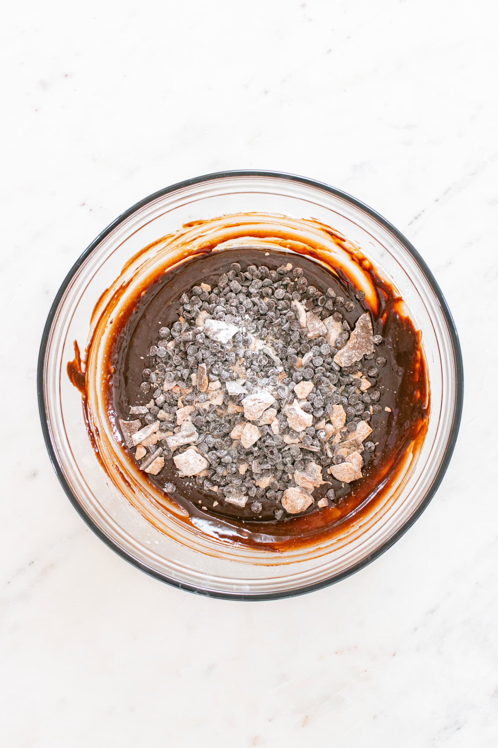A glass bowl containing brownie batter, chocolate chips, and unincorporated dry ingredients sits on a white marble surface.