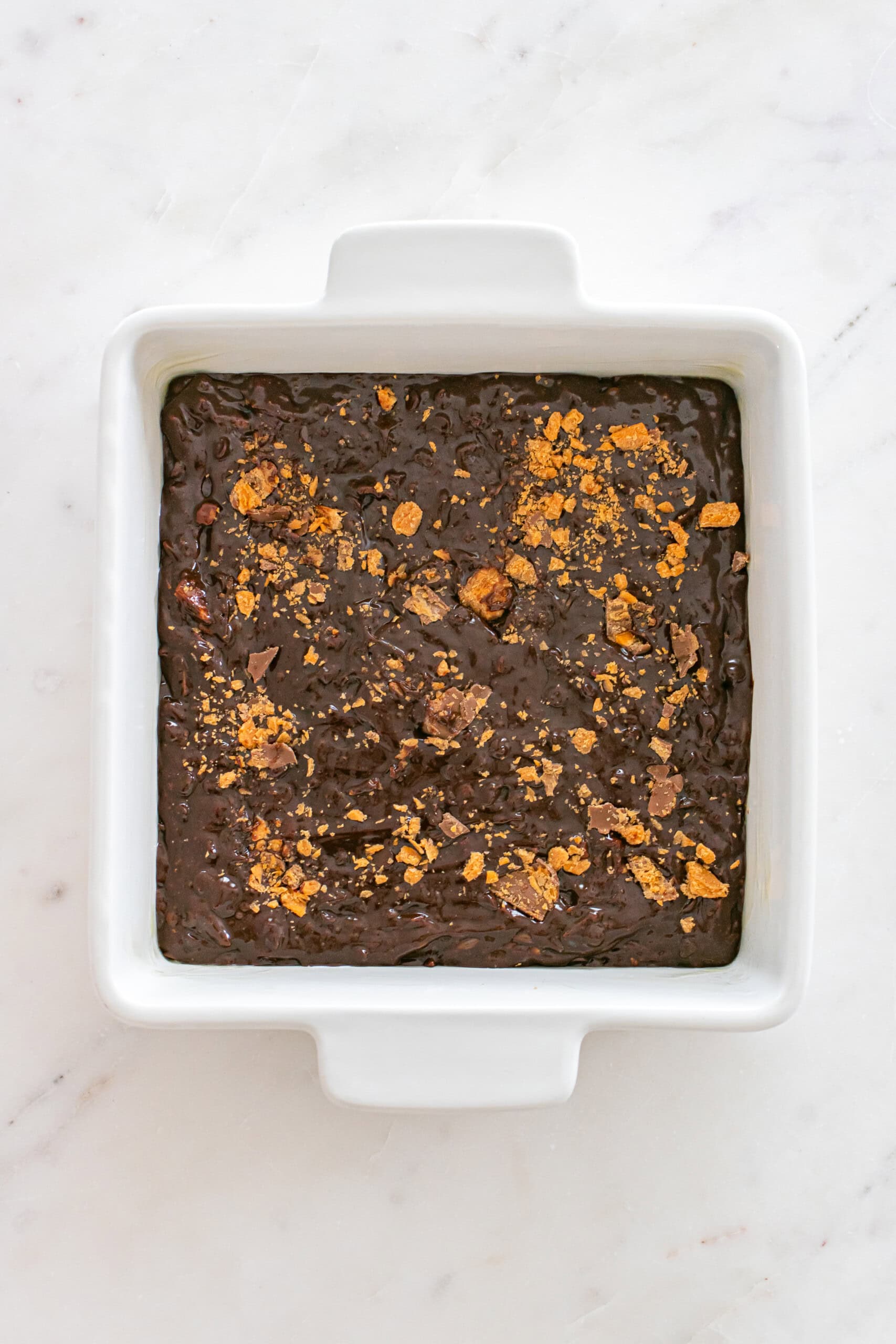 A square white baking dish filled with unbaked chocolate batter, topped with crumbled pieces, sits on a light marble surface.