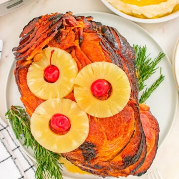 A crockpot ham garnished with pineapple rings and cherries, served on a plate with sprigs of rosemary, next to a bowl of mashed potatoes.