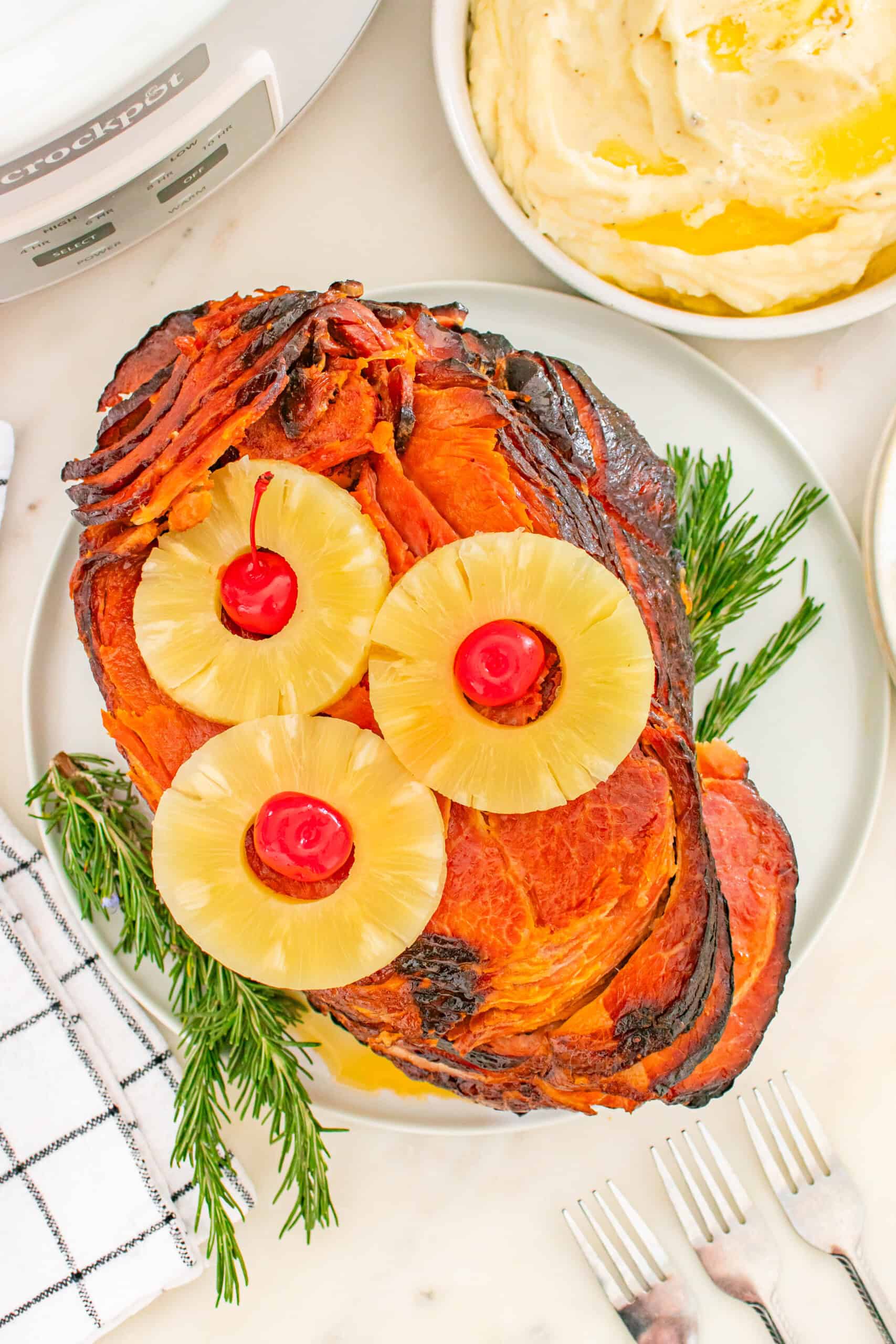 A crockpot ham garnished with pineapple rings and cherries, served on a plate with sprigs of rosemary, next to a bowl of mashed potatoes.