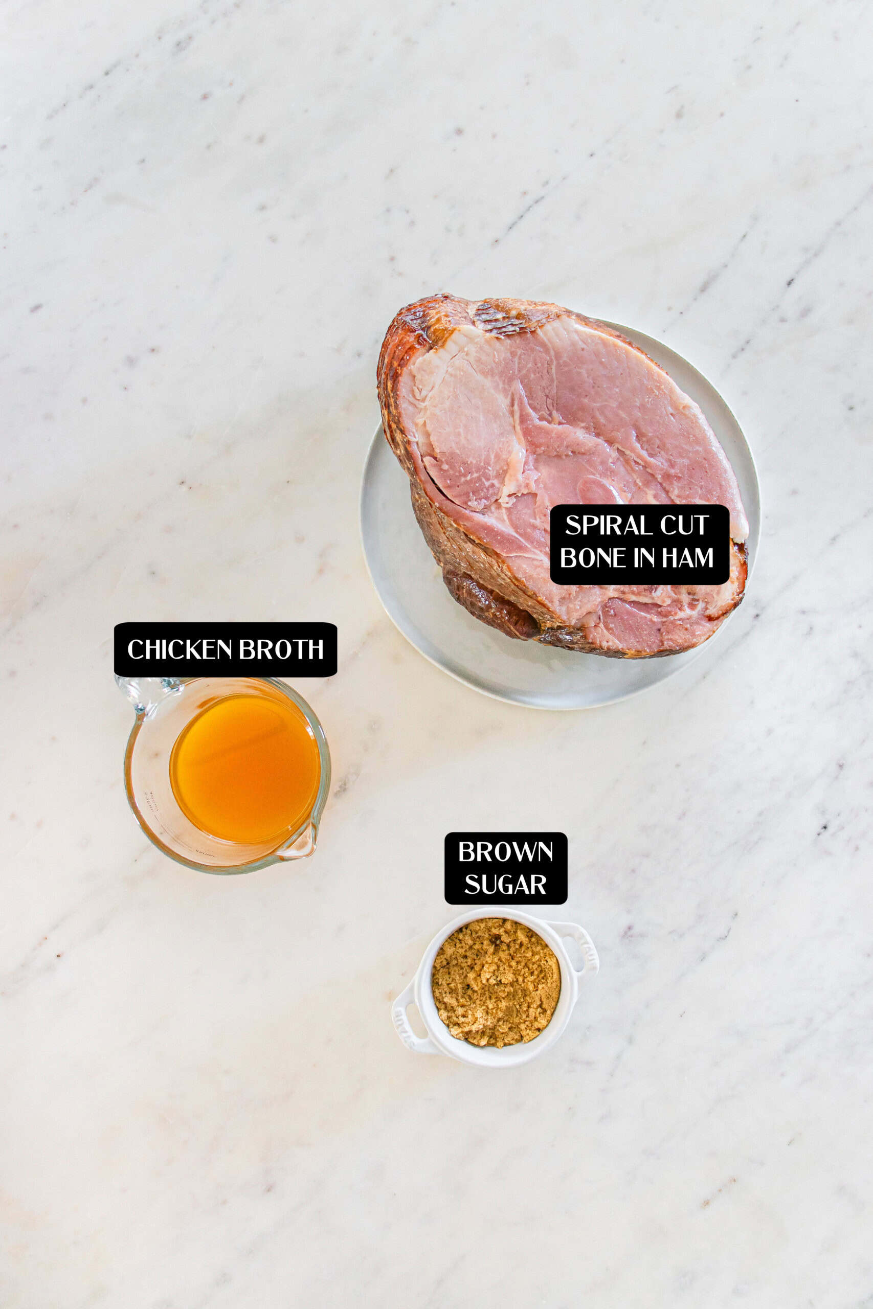 A bowl with spiral cut bone-in ham, a measuring cup of chicken broth, and a small bowl of brown sugar on a light surface—perfect ingredients for making crockpot ham—each labeled with text.