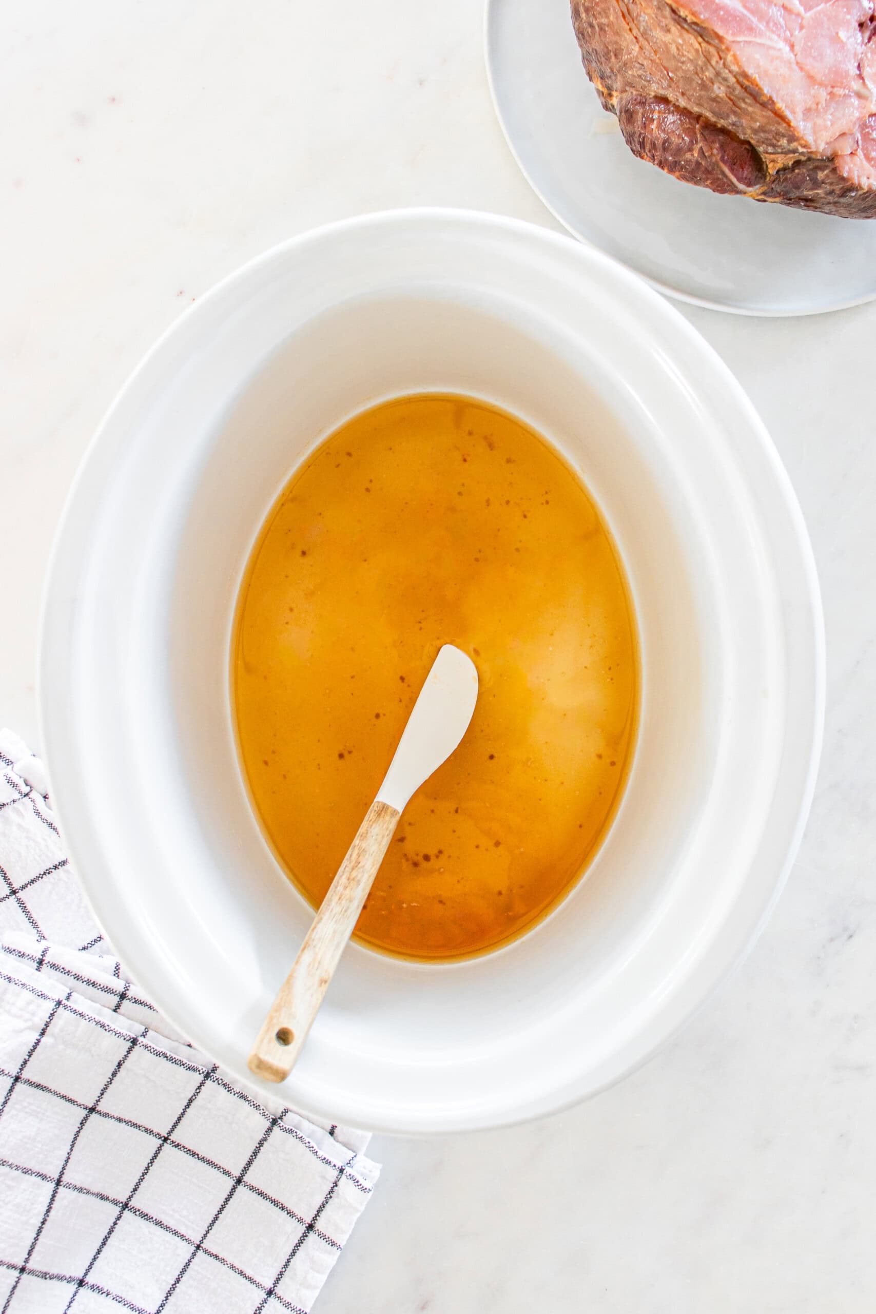 White slow cooker filled with brown liquid and a white spatula inside, showcasing crockpot ham on a plate beside a white towel with black stripes.