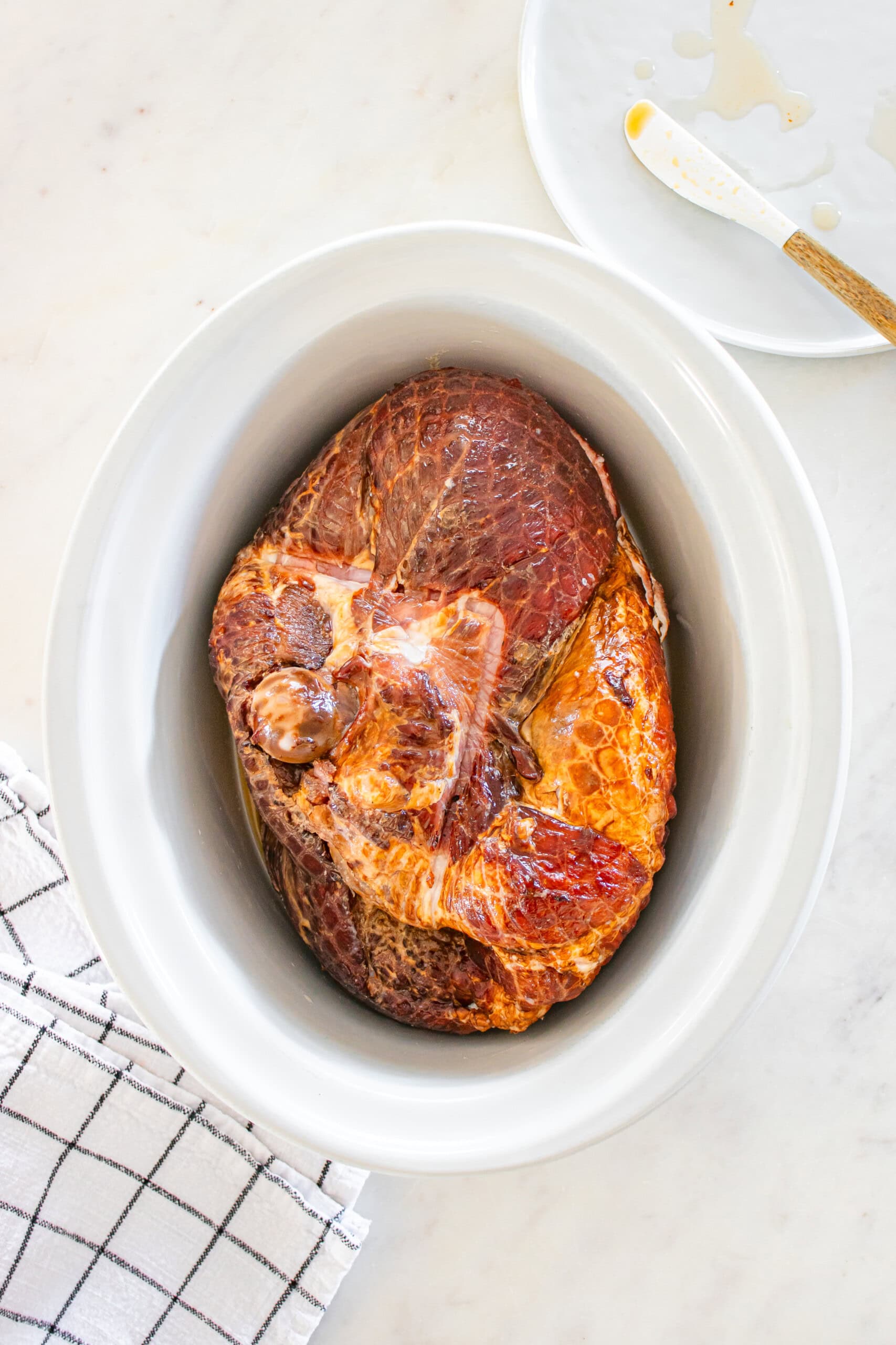 A crockpot ham sits in a white slow cooker on a marble countertop, next to a checkered towel and a plate with a spreading knife.