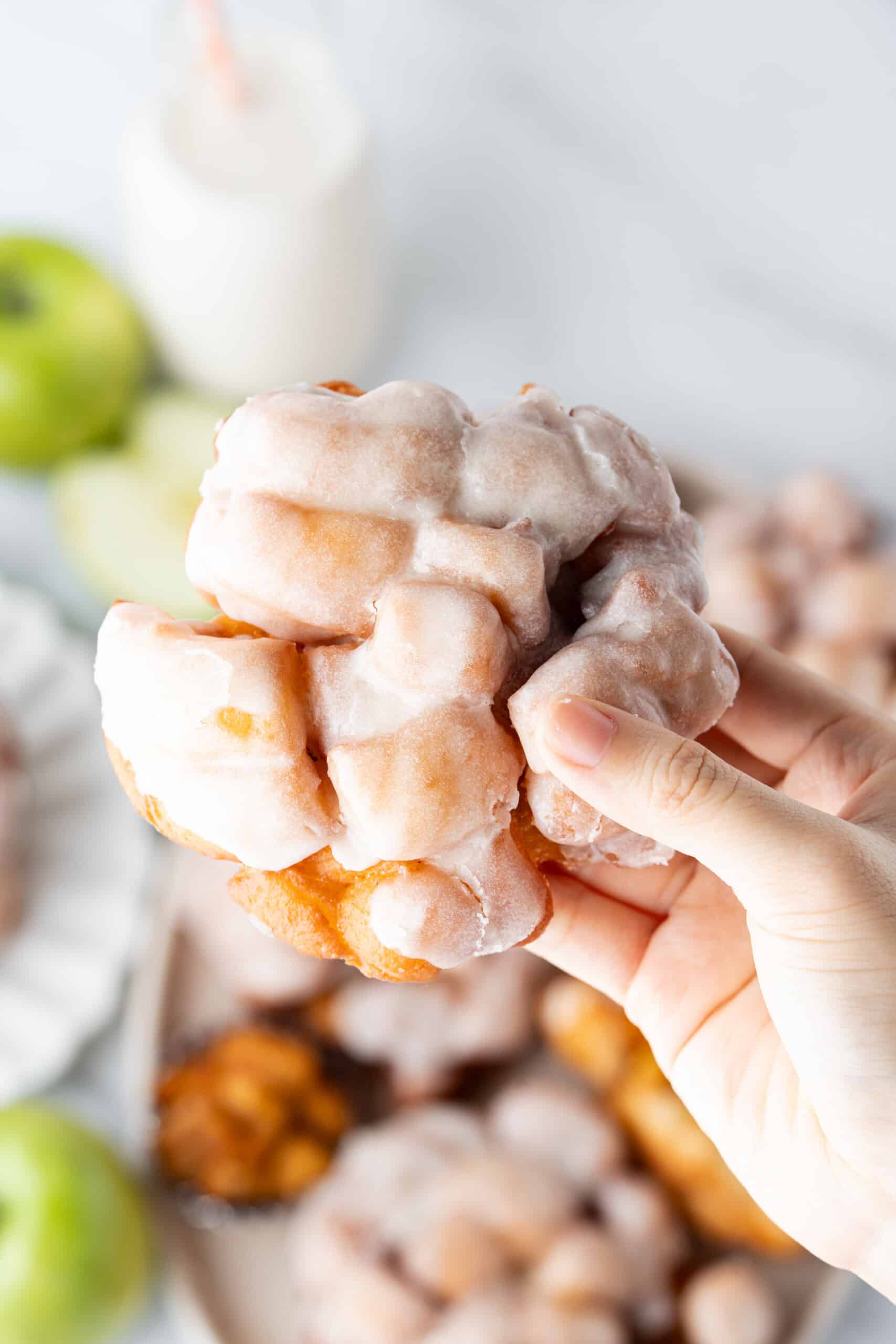 Apple fritter covered in sweet glaze and held by hand with fresh apples in the background.