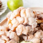 Glazed apple fritters stacked on a serving platter with green apples.