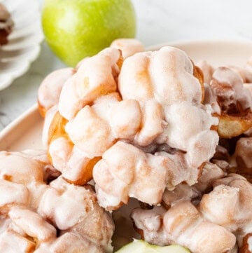 Glazed apple fritters stacked on a serving platter with green apples.