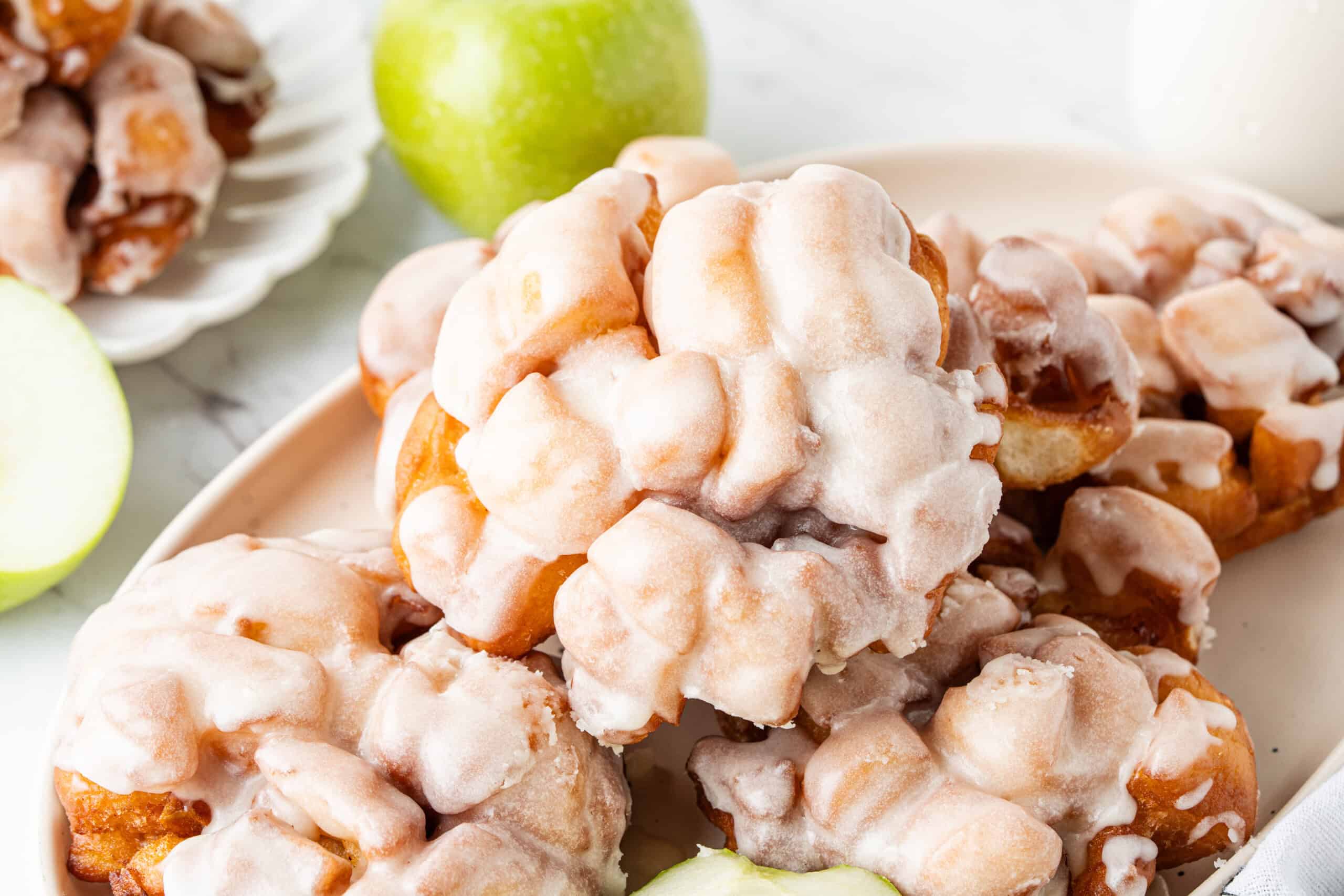 Glazed apple fritters stacked on a serving platter with green apples.