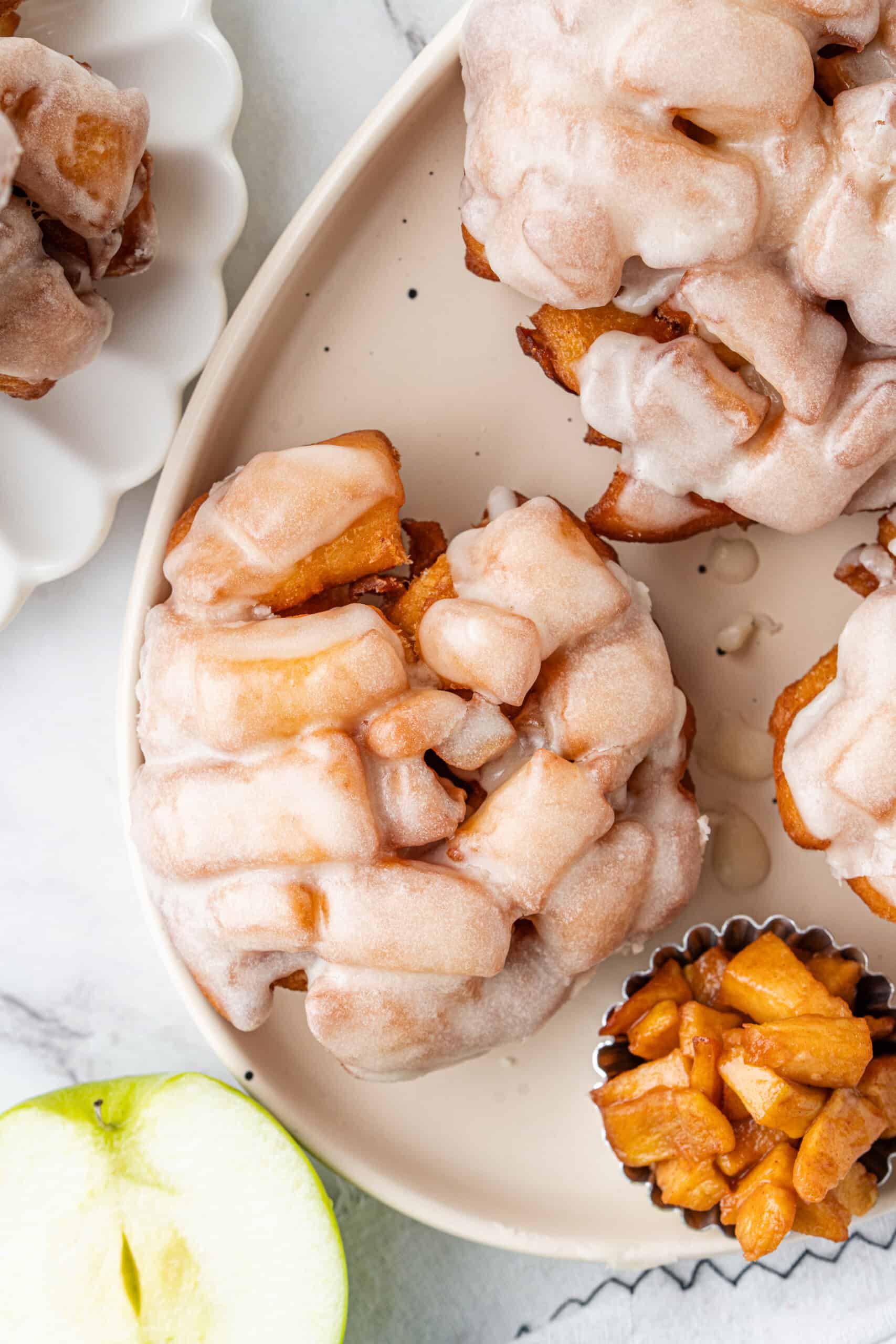 Glazed apple fritters on a serving platter with cooked cinnamon apples and sliced green apple.