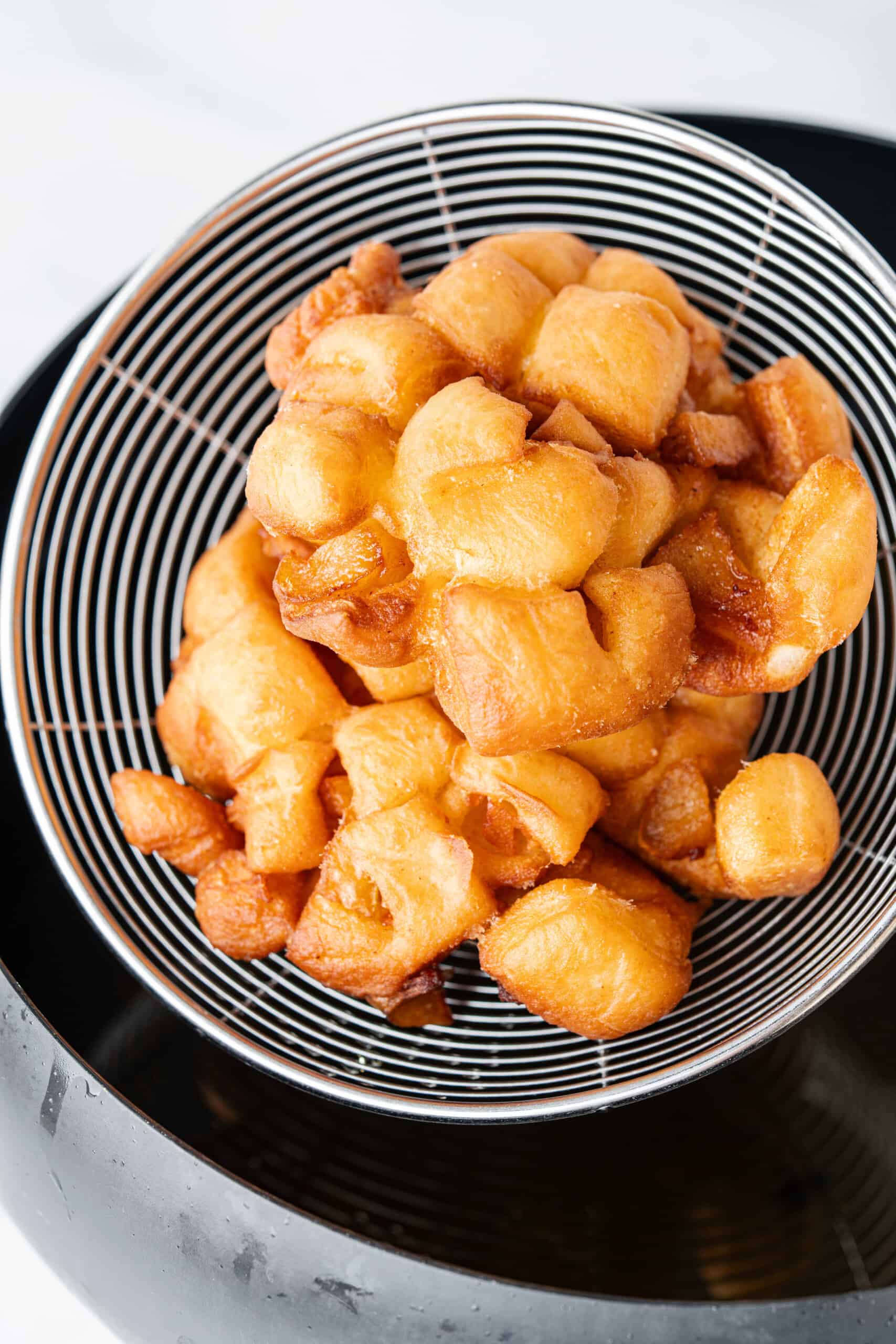 Freshly fried apple fritters draining in a wire skimmer over hot oil.
