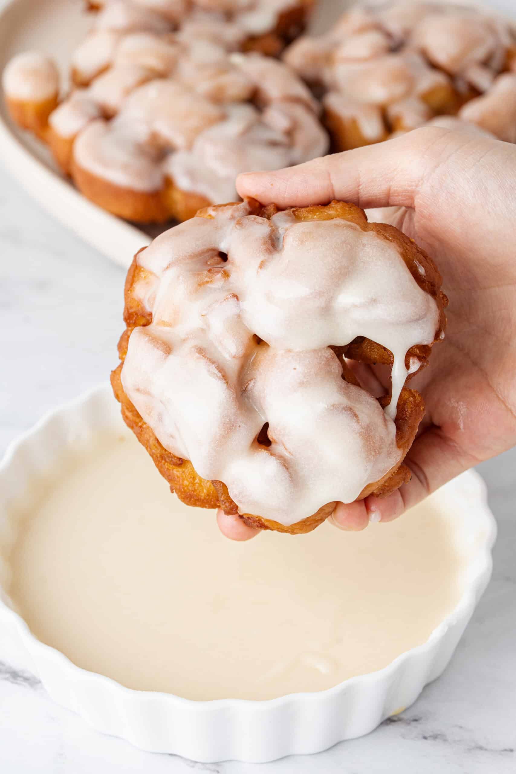 A glazed apple fritter held above a bowl of glaze.