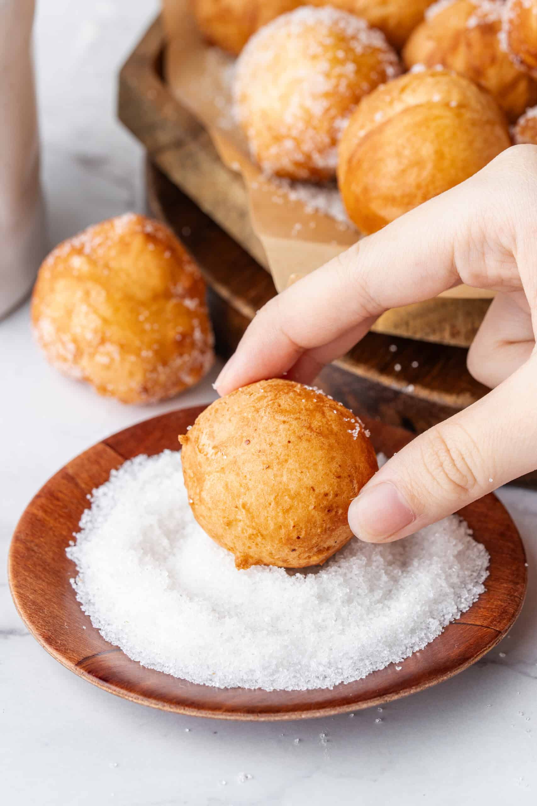 Hand rolling a freshly fried donut hole in sugar on a small wooden plate.