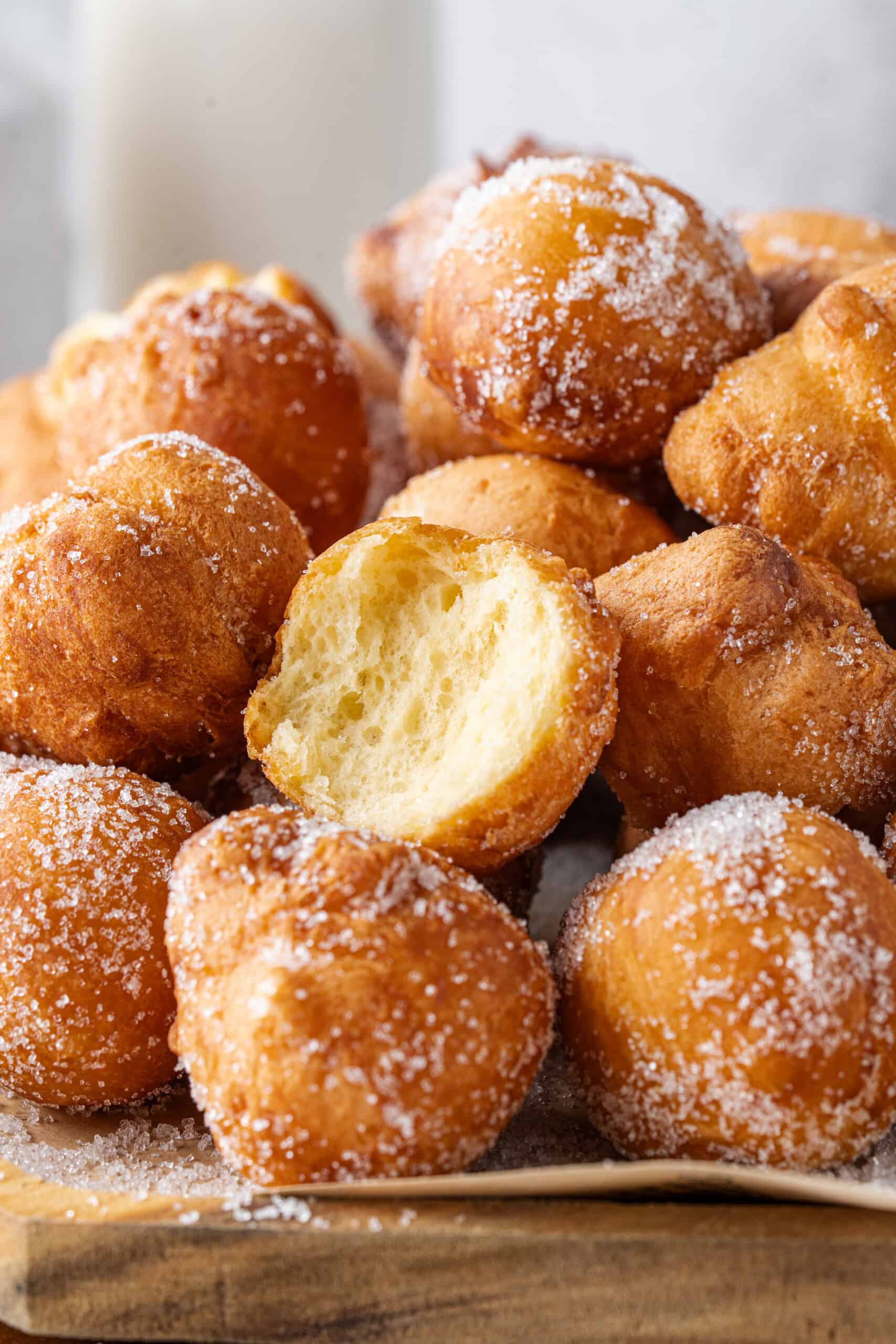 Pile of sugar-coated donut holes on a wooden board, with one donut hole cut open to show its fluffy inside.