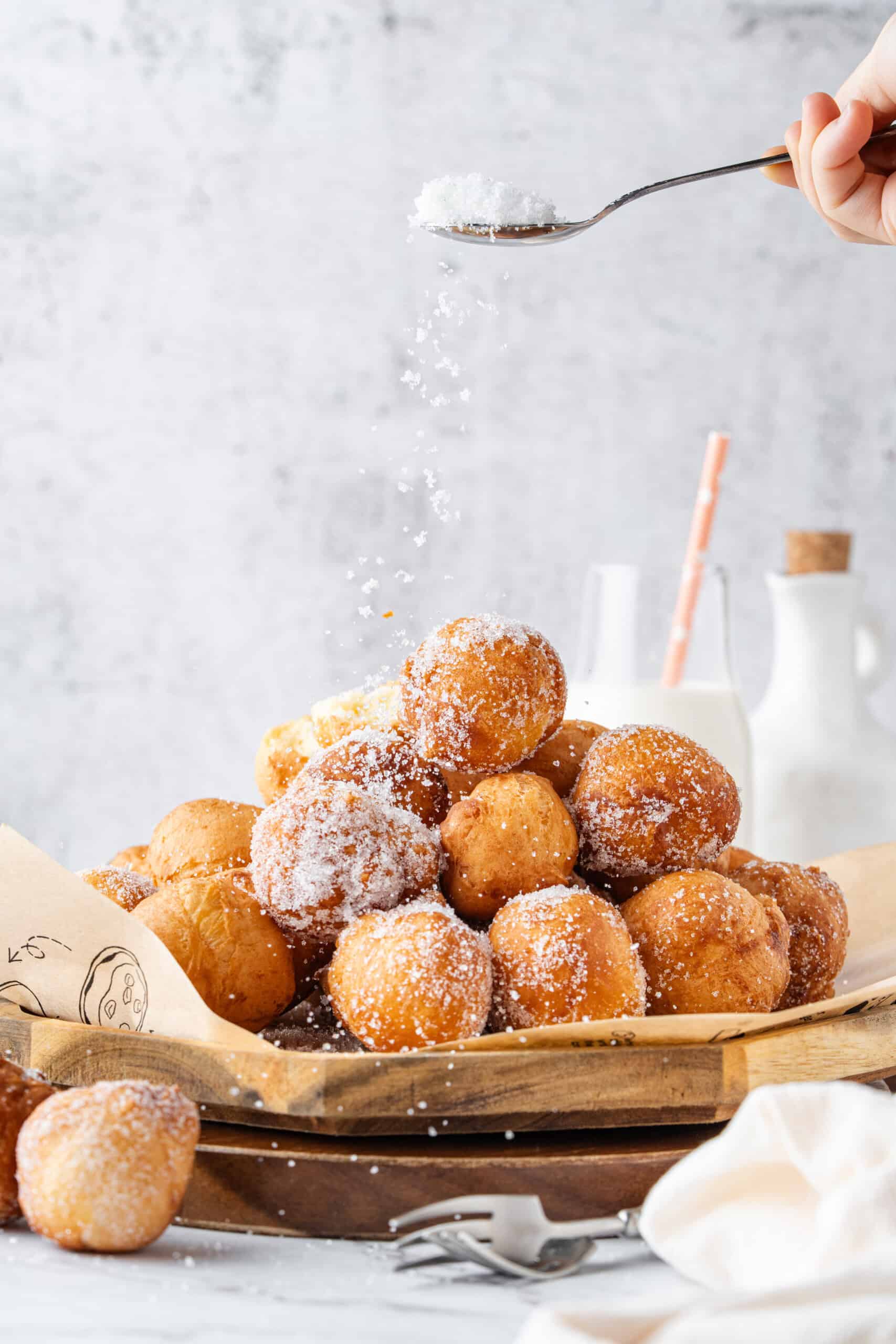 A spoon sprinkles sugar over a pile of sugar-coated donut holes on a wooden tray, with milk in the background.