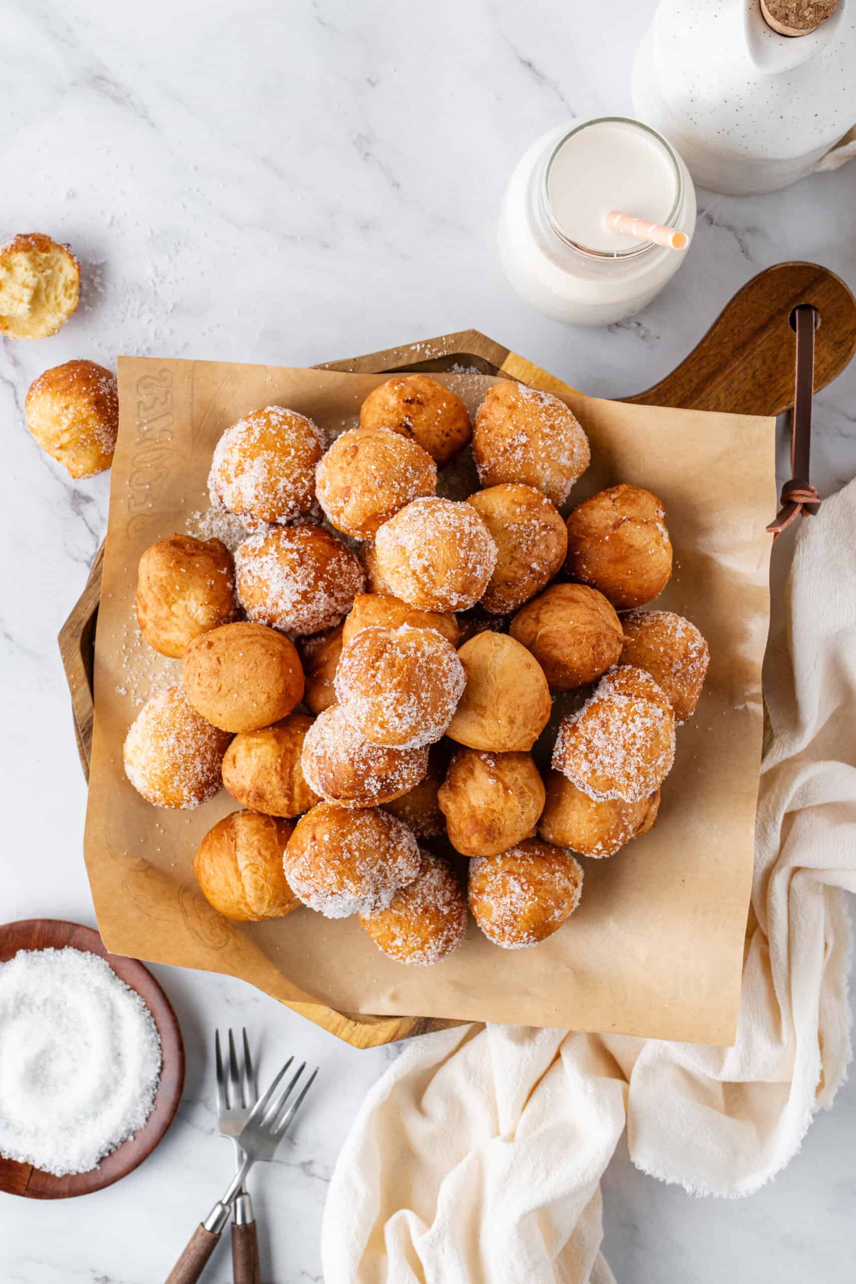 A tray of sugar-coated donut holes on parchment paper, served with a bottle of milk on a marble countertop.
