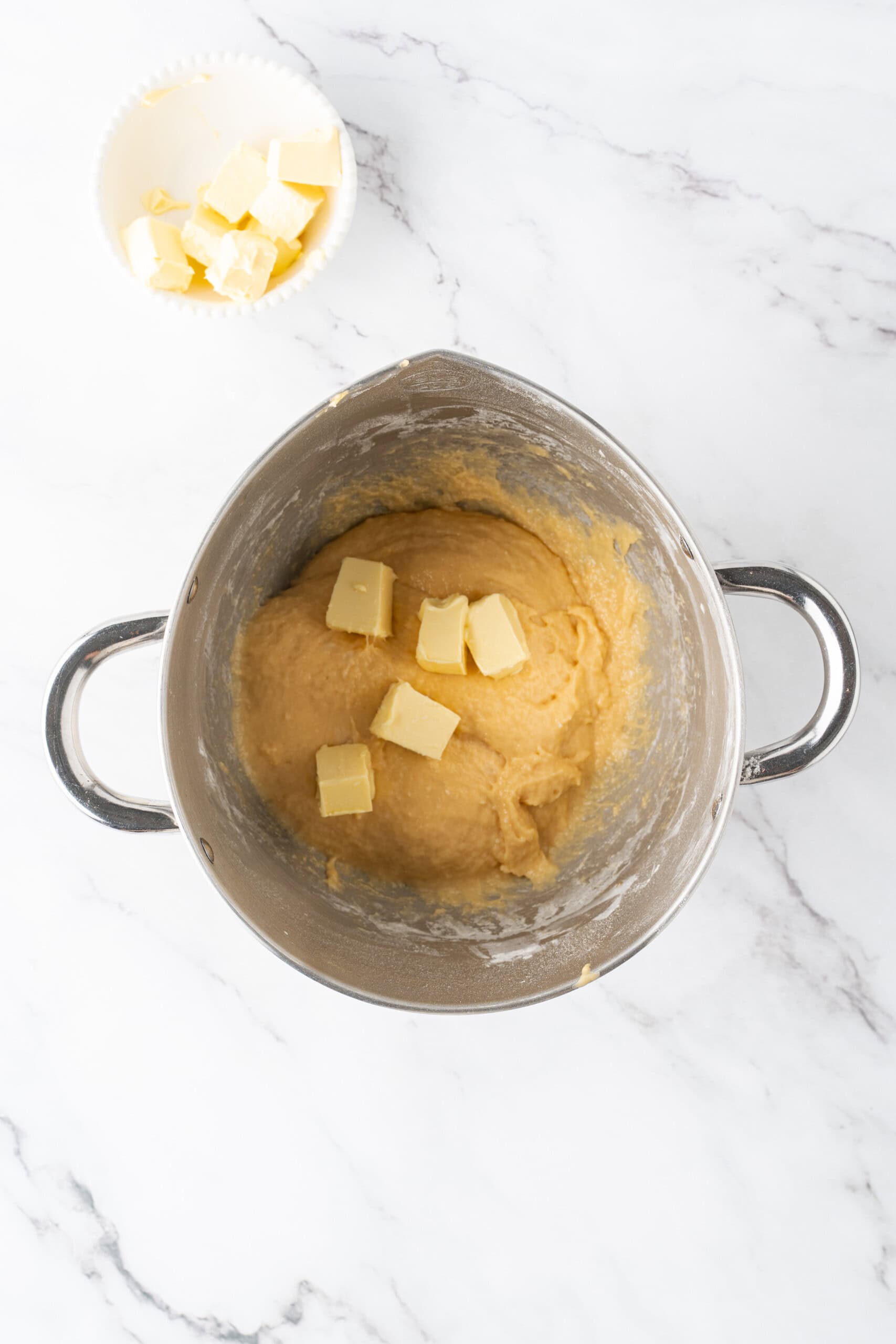 Butter cubes being added to a bowl of dough in a mixing bowl on a marble countertop.