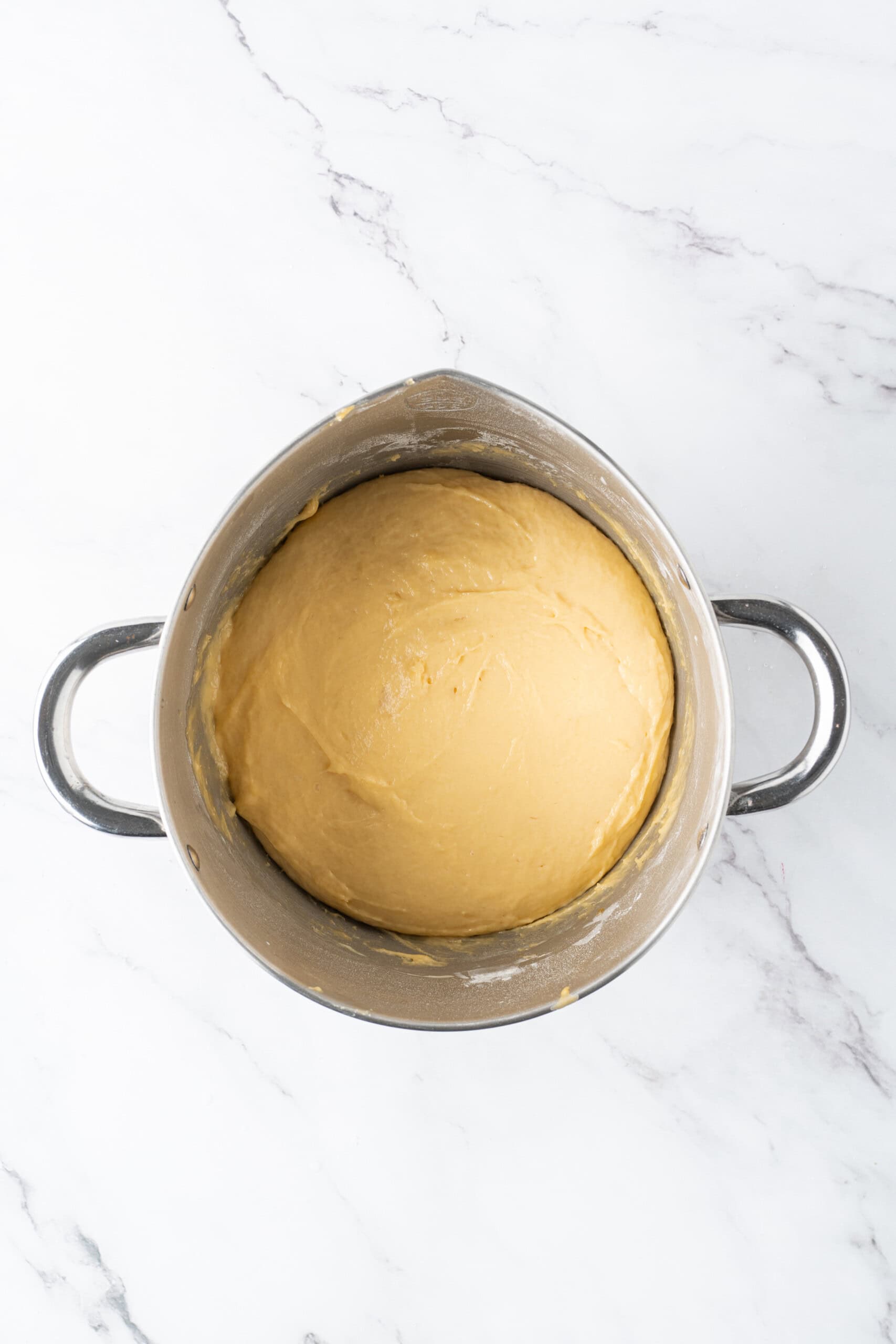 Risen dough doubled in size in a metal mixing bowl on a marble countertop.