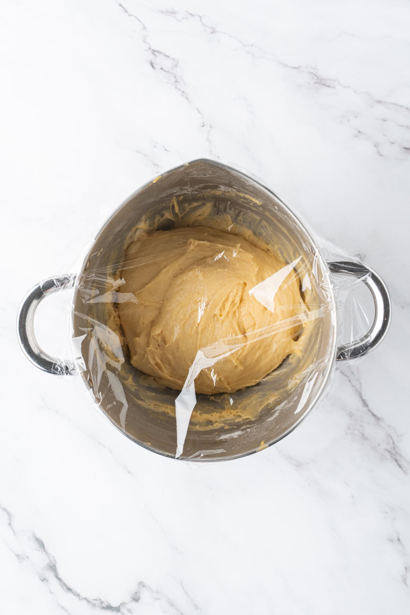 Dough covered with plastic wrap in a metal mixing bowl, ready to rise.