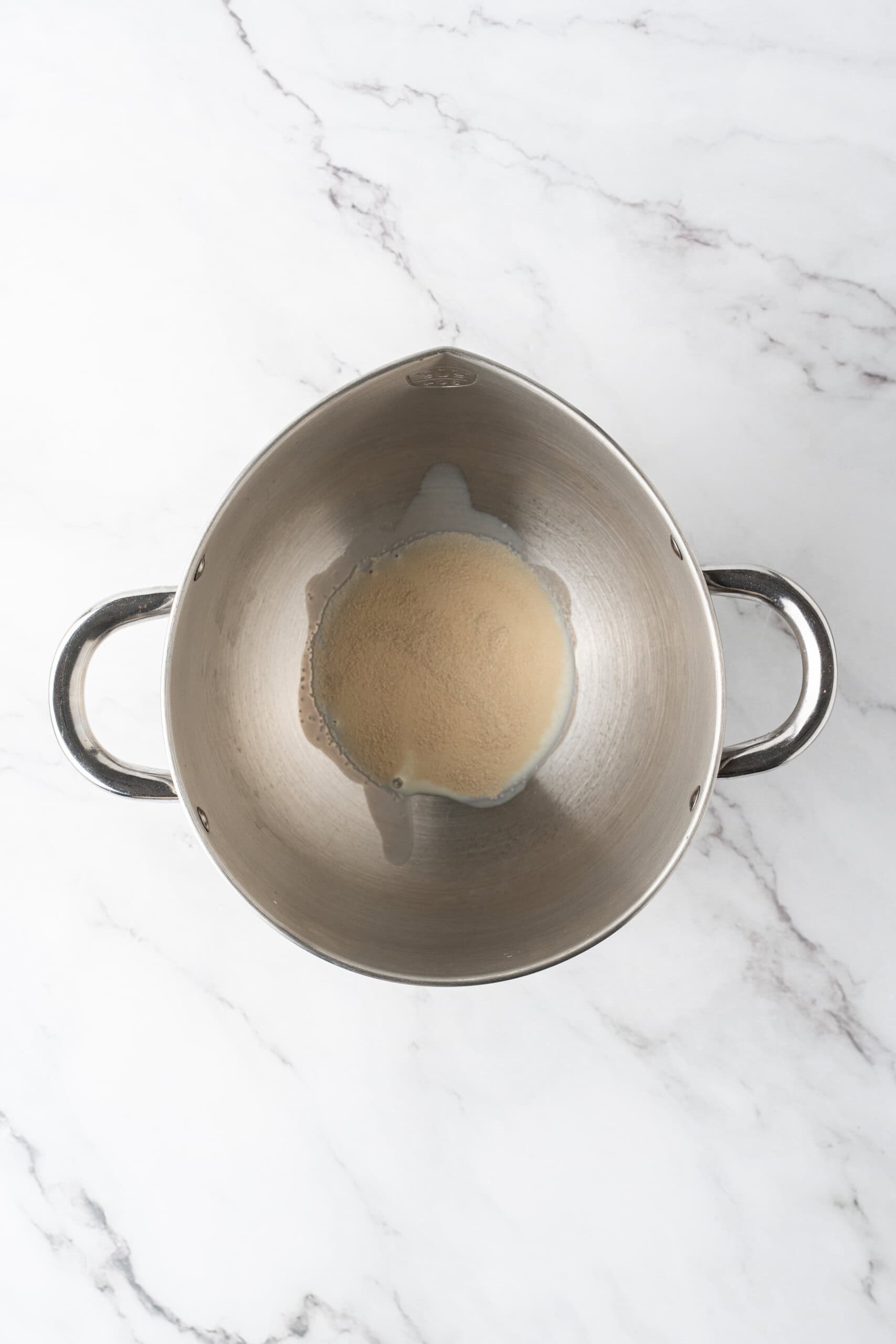 Yeast and milk in a metal mixing bowl on a marble countertop