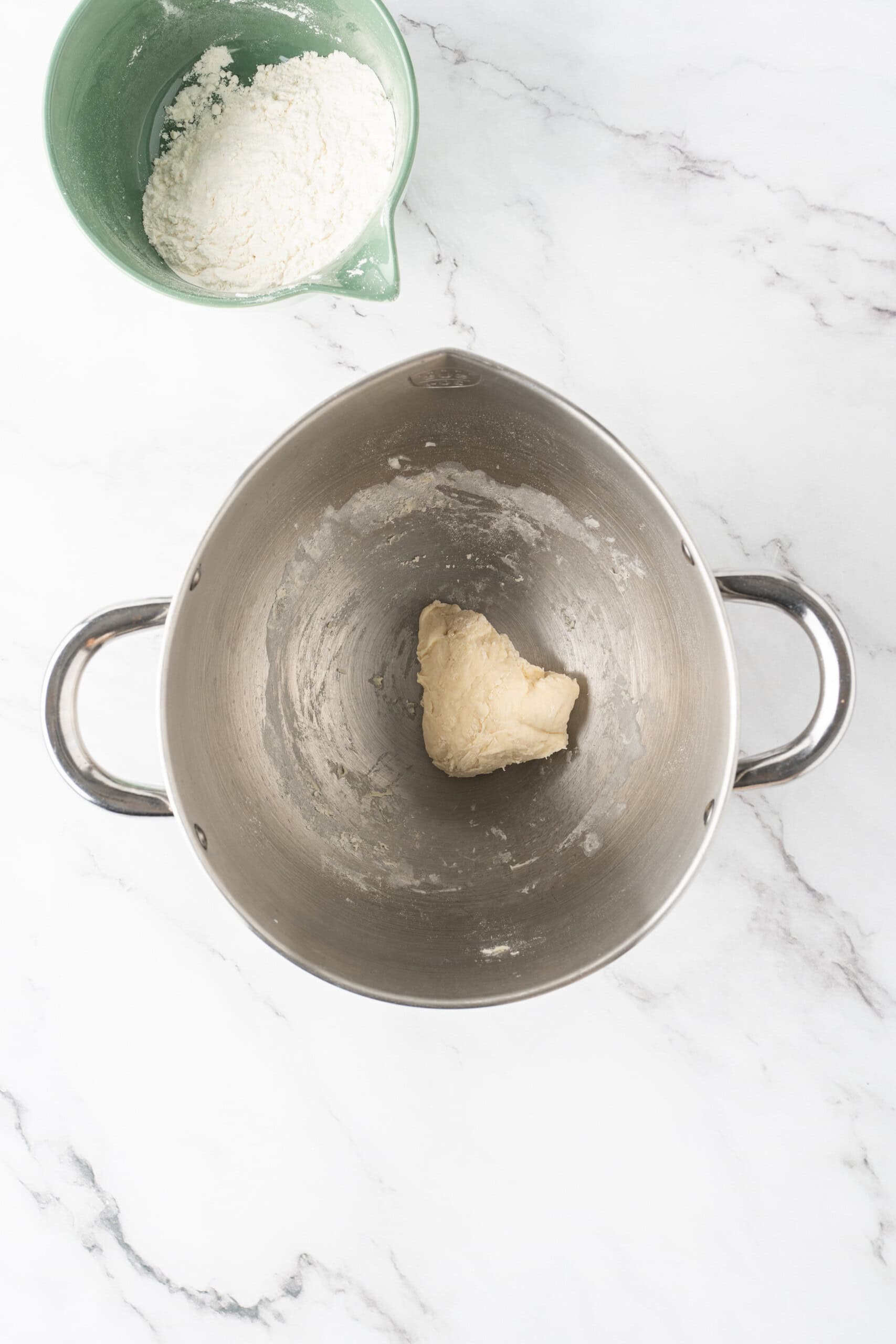 Partially mixed donut dough in a metal mixing bowl with extra bread flour nearby