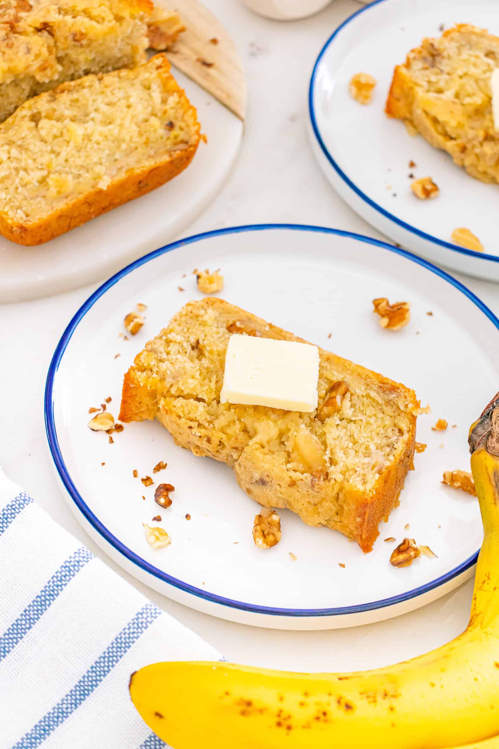 Banana bread slices topped with butter, served on plates alongside fresh bananas and chopped walnuts.