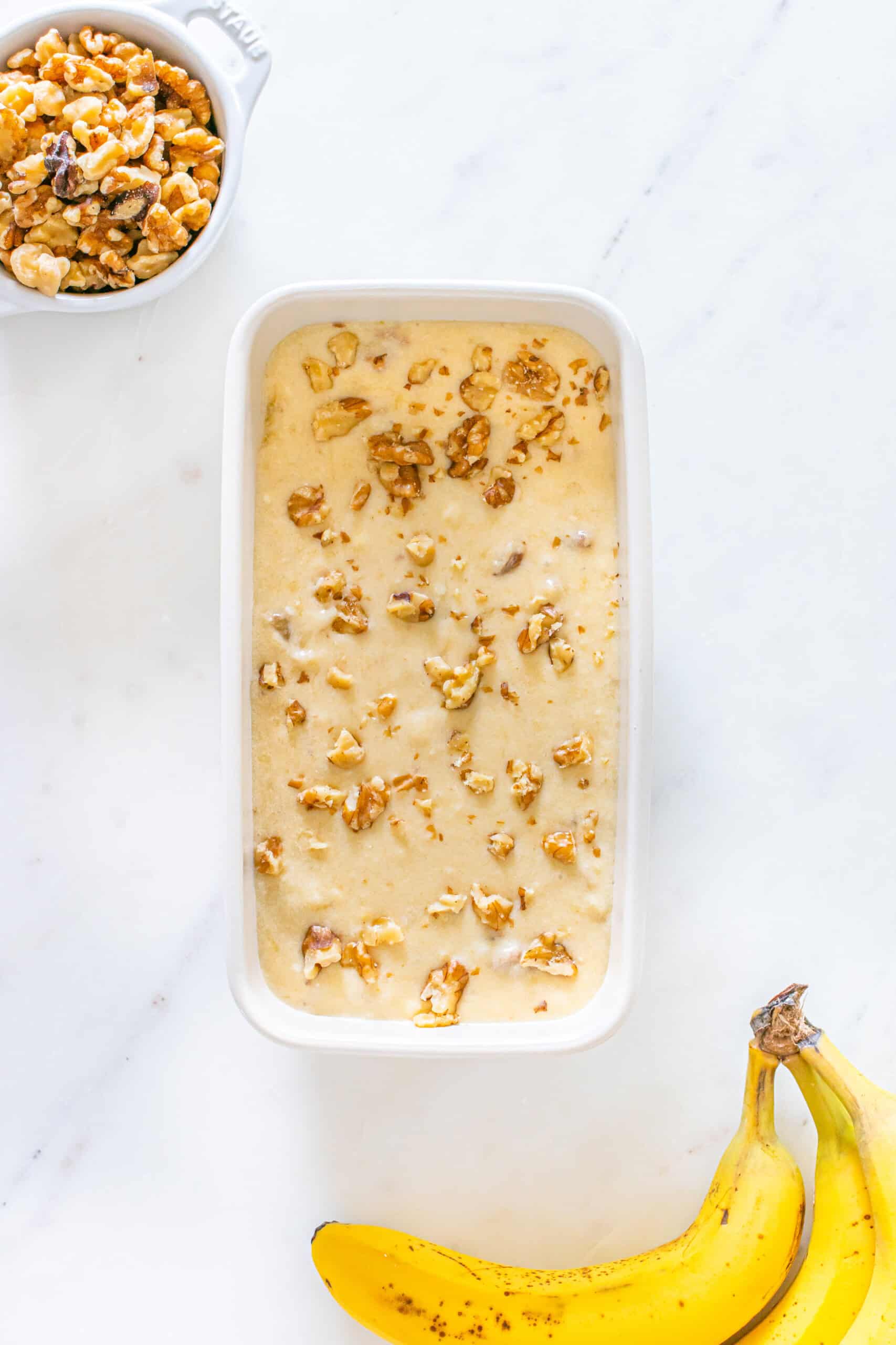 Banana bread batter topped with chopped walnuts, ready for baking, with fresh bananas nearby.