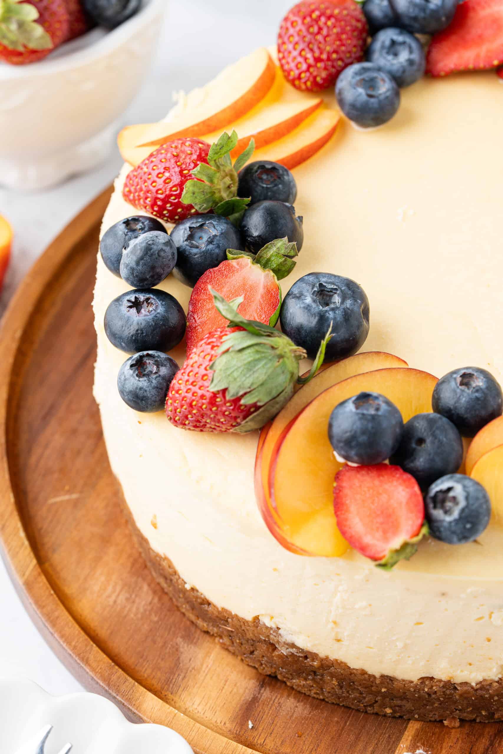 Close-up of a creamy baked cheesecake with a graham cracker crust, decorated with fresh strawberries, blueberries, and peach slices.