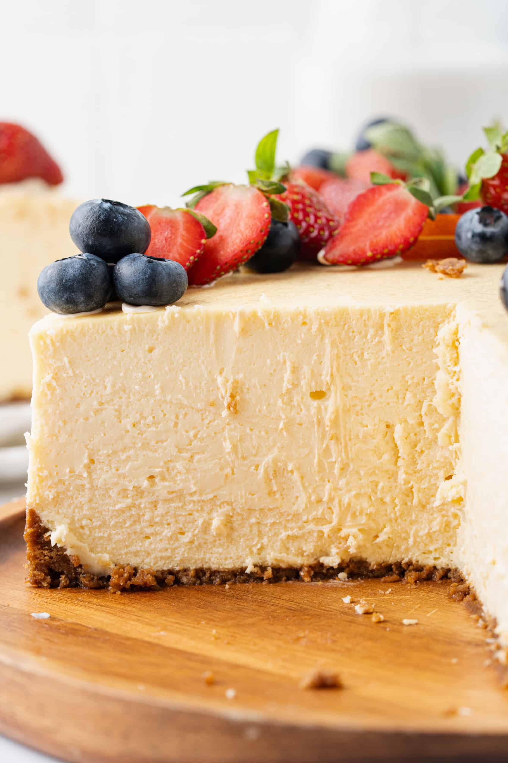 Close-up of a rich and creamy baked cheesecake showing its smooth texture and thick graham cracker crust, topped with fresh strawberries and blueberries on a wooden serving board.