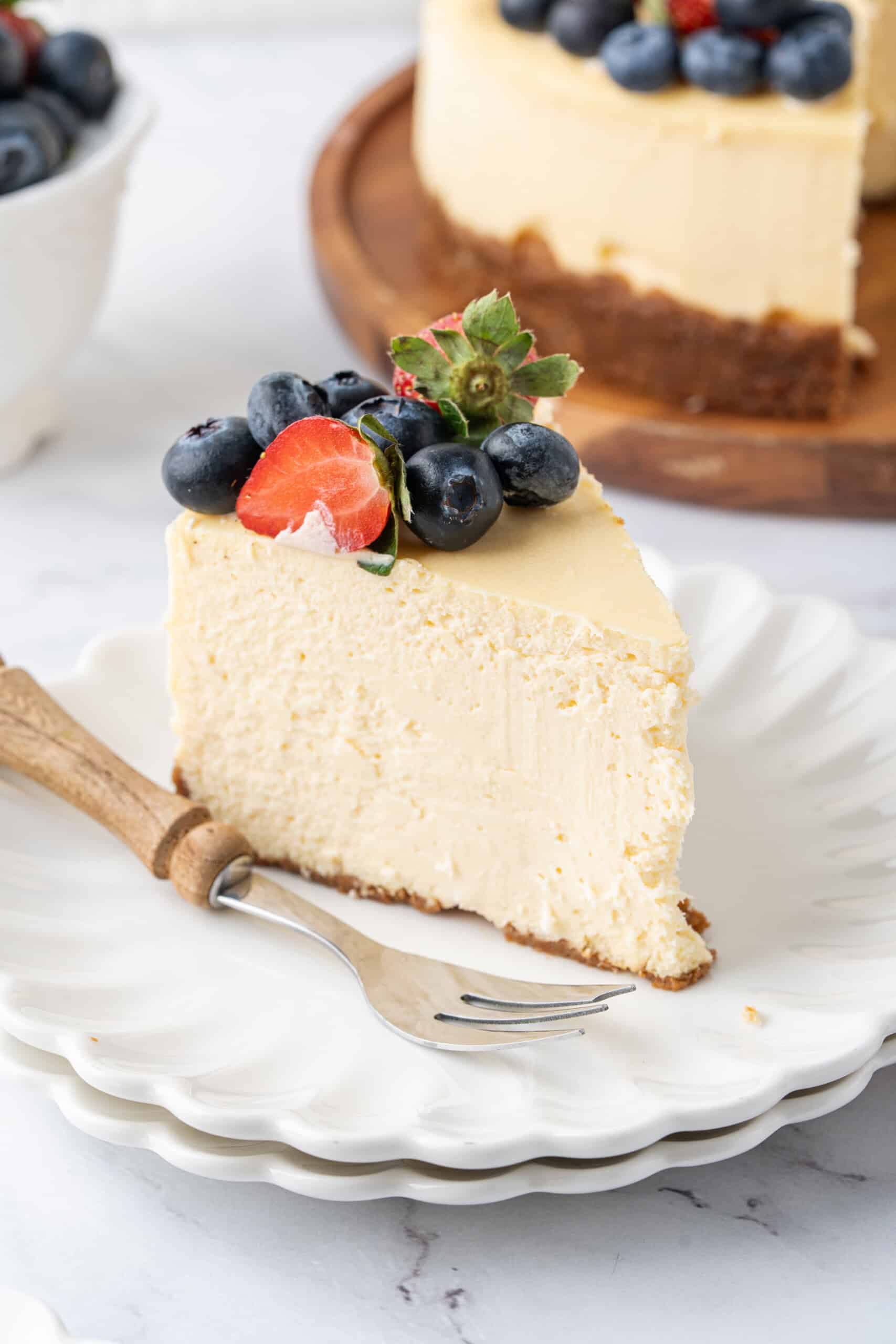 Slice of creamy baked cheesecake with a graham cracker crust, topped with fresh strawberries and blueberries, served on a white plate with the remaining cheesecake in the background.