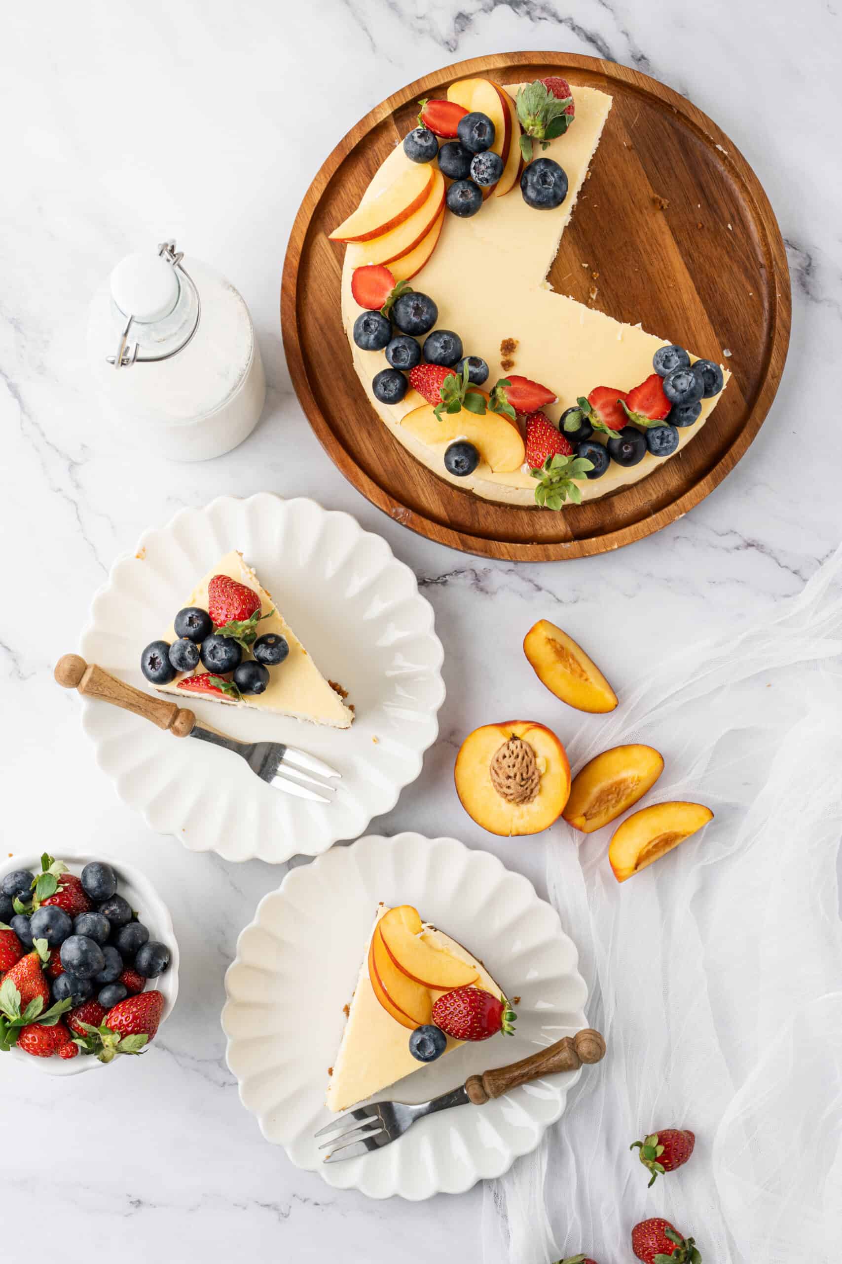 Overhead view of a baked cheesecake topped with fresh strawberries, blueberries, and peach slices, with two plated slices ready to serve.