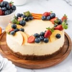 Whole baked cheesecake with a buttery graham cracker crust, decorated with fresh strawberries, blueberries, and peach slices on a wooden serving board.
