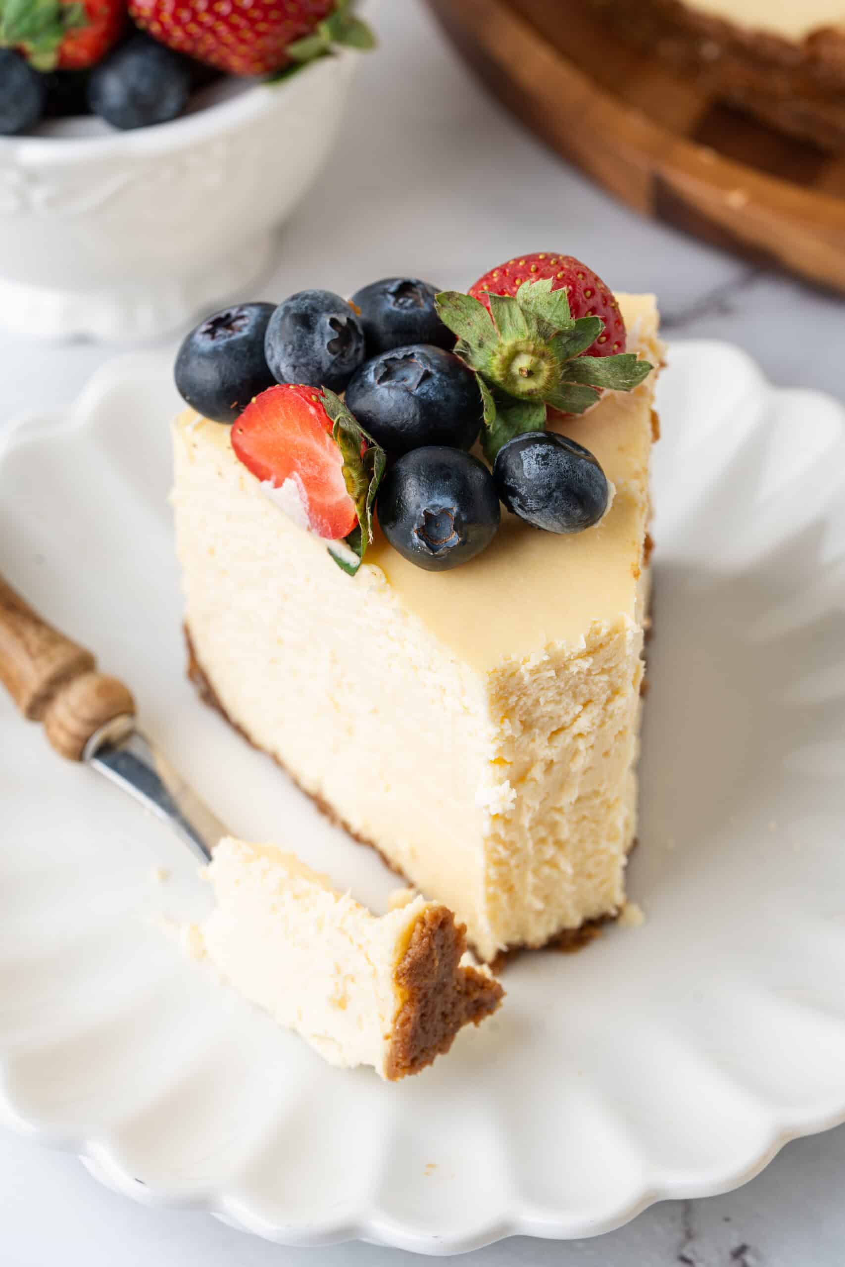 Close-up of a creamy baked cheesecake slice with a graham cracker crust, topped with fresh strawberries and blueberries, with a bite on the fork ready to enjoy.