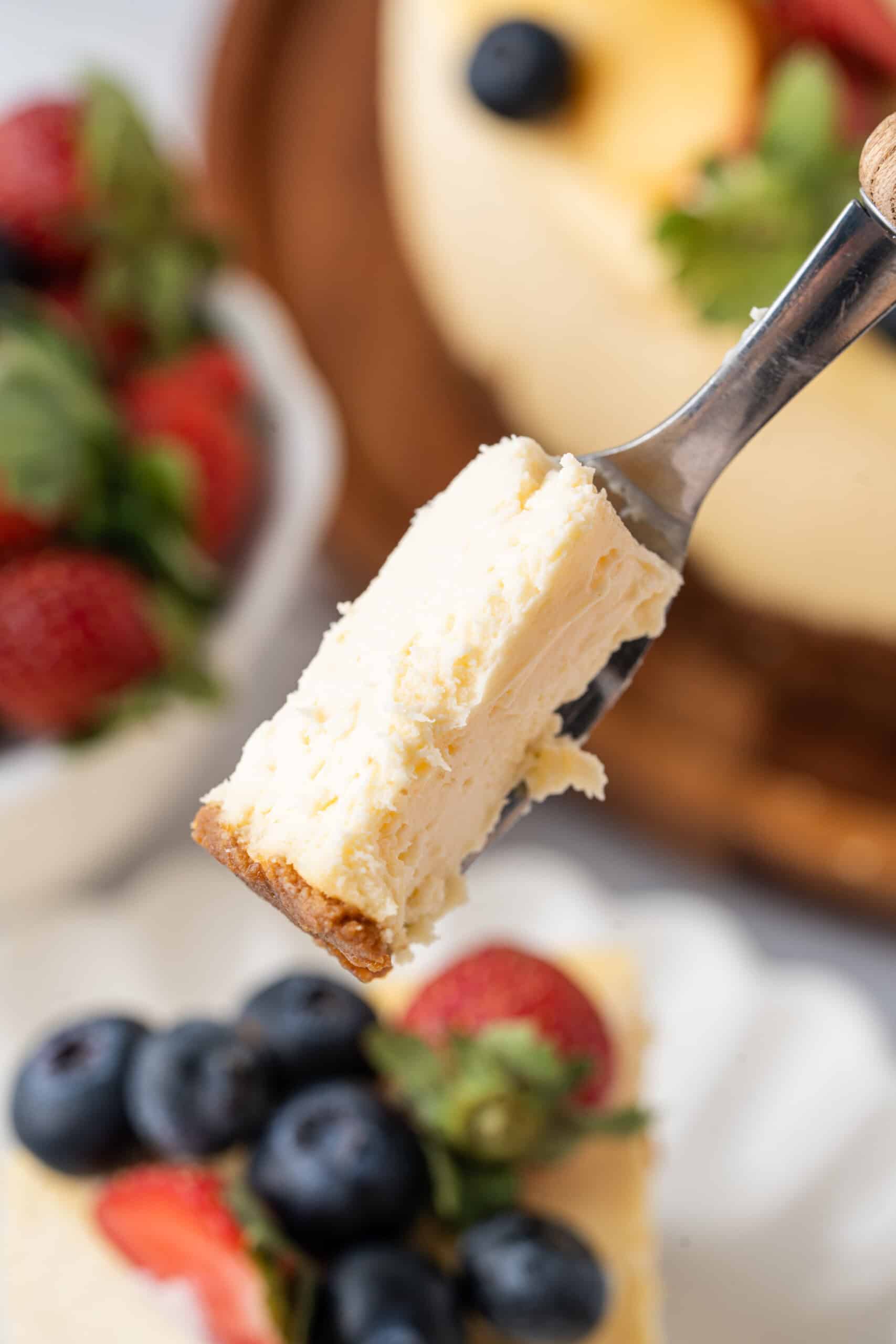 Close-up of a fork holding a bite of creamy baked cheesecake with a graham cracker crust, with fresh berries and the cheesecake softly blurred in the background.