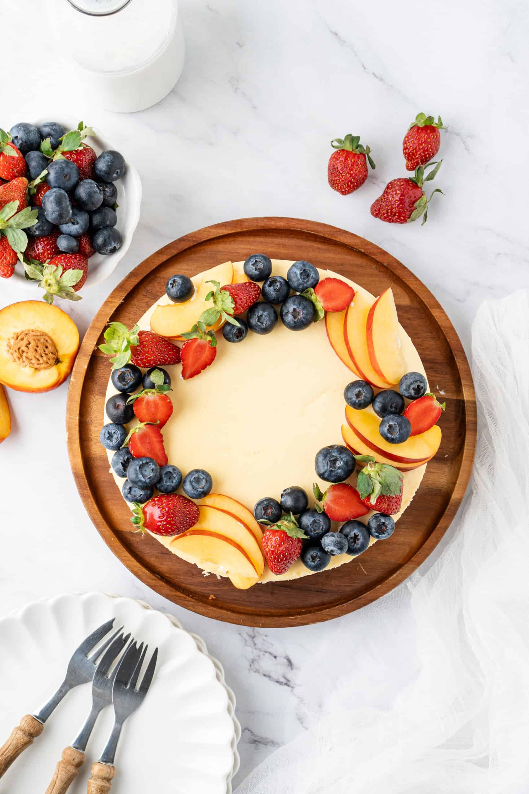 Overhead view of a whole baked cheesecake decorated with fresh strawberries, blueberries, and peach slices on a wooden serving board, ready to slice and serve.