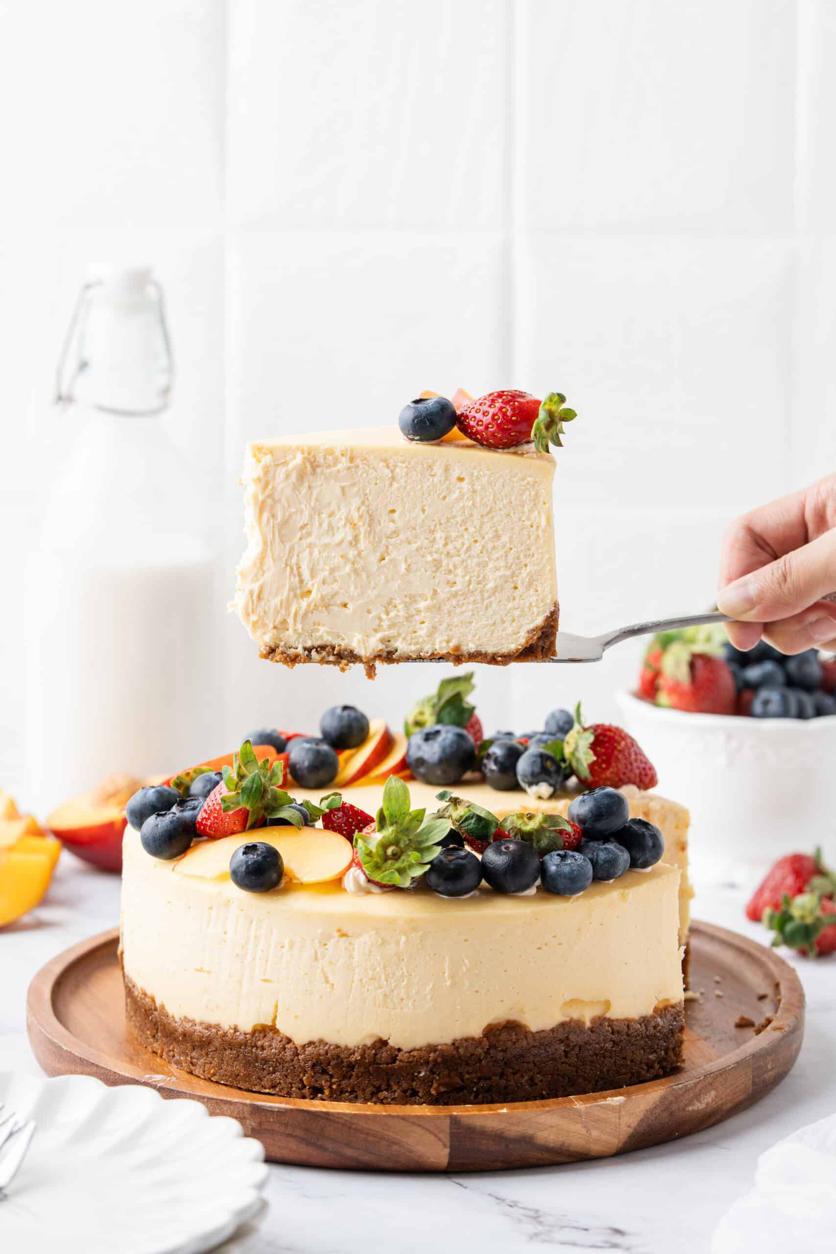 Hand holding a slice of classic cheesecake with graham cracker crust topped with fresh strawberries and blueberries.