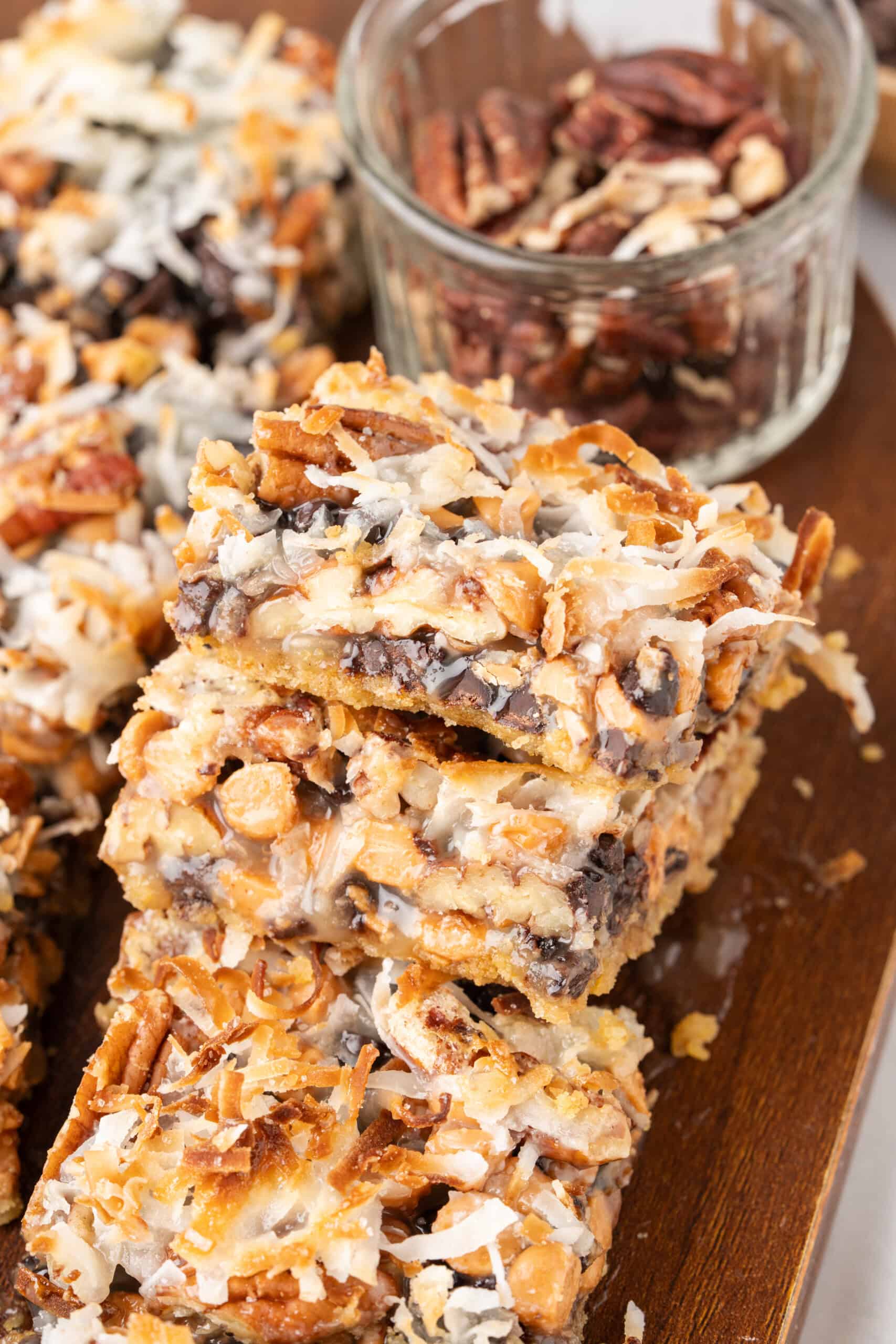 Stacked Hello Dolly Bars with chocolate chips, butterscotch chips, coconut, and pecans on a wooden board.