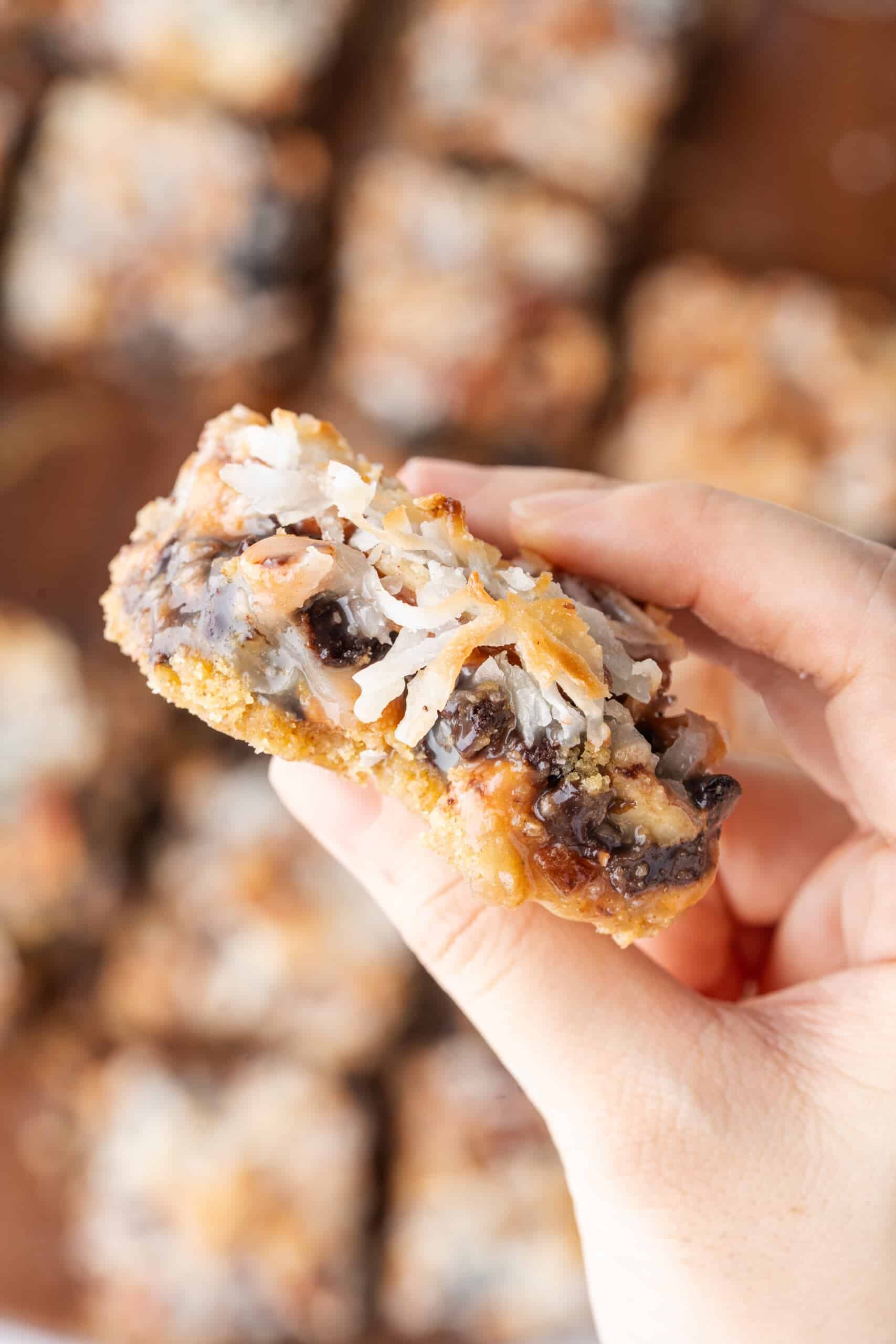 Hand holding a 7-Layer Bars with chocolate chips, butterscotch chips, coconut, and pecans.