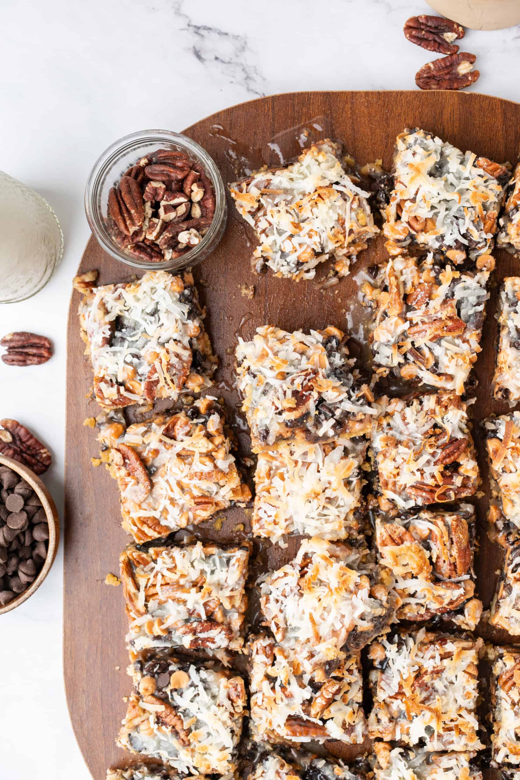Overhead view of homemade magic bars topped with coconut, pecans, chocolate chips, and butterscotch chips on a wooden board.