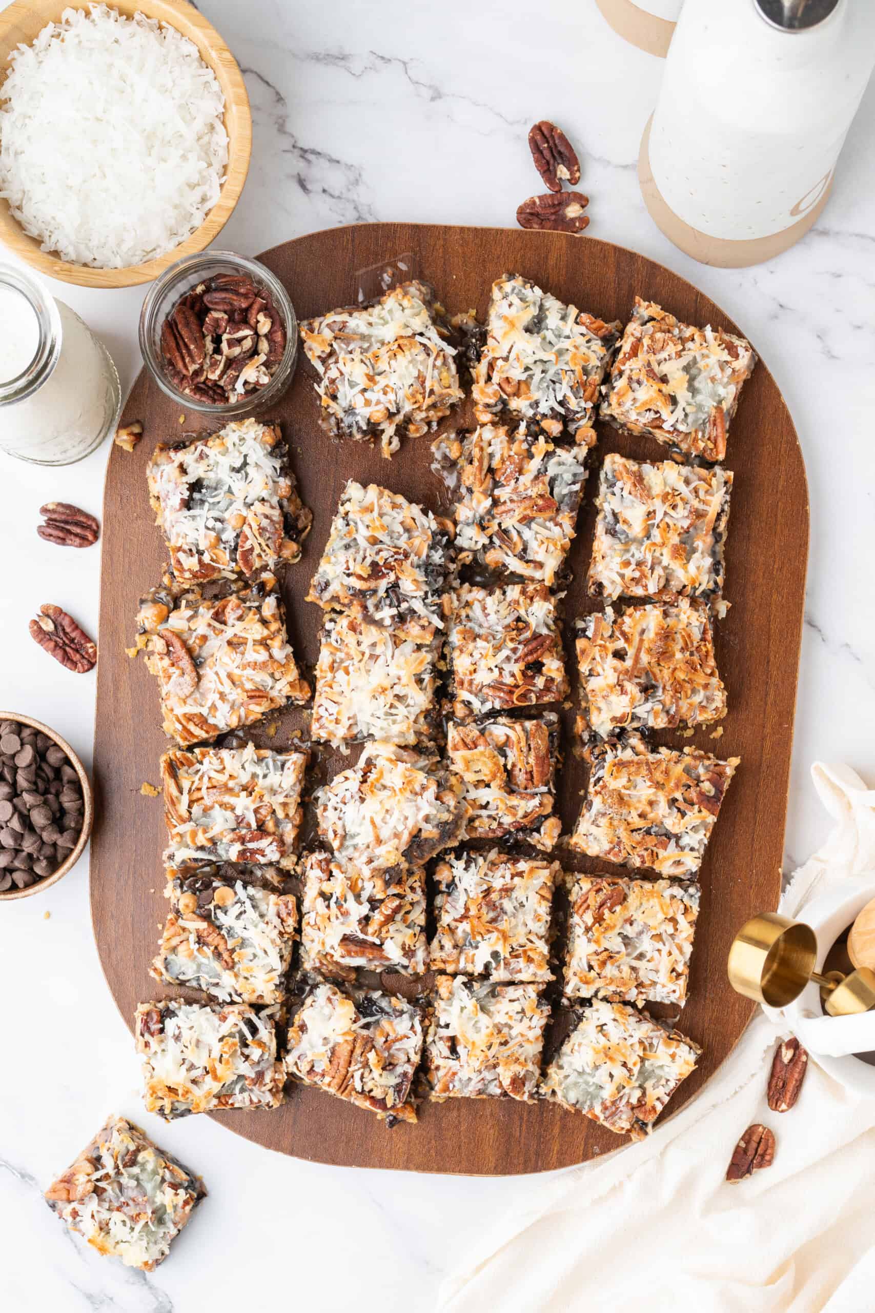 Overhead view of homemade magic bars topped with coconut, pecans, chocolate chips, and butterscotch chips on a wooden board.