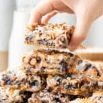 Hand holding a magic bar with chocolate chips, coconut, and pecans above a stack of magic bars.