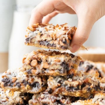 Hand holding a magic bar with chocolate chips, coconut, and pecans above a stack of magic bars.