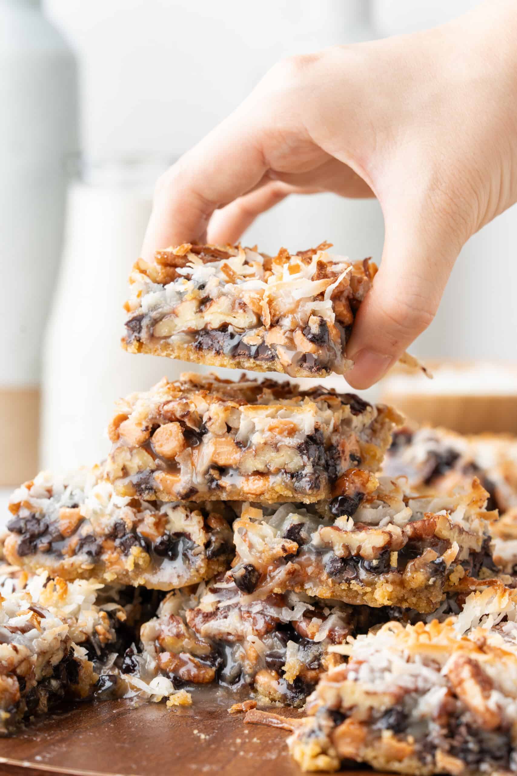 Hand holding a magic bar with chocolate chips, coconut, and pecans above a stack of magic bars.